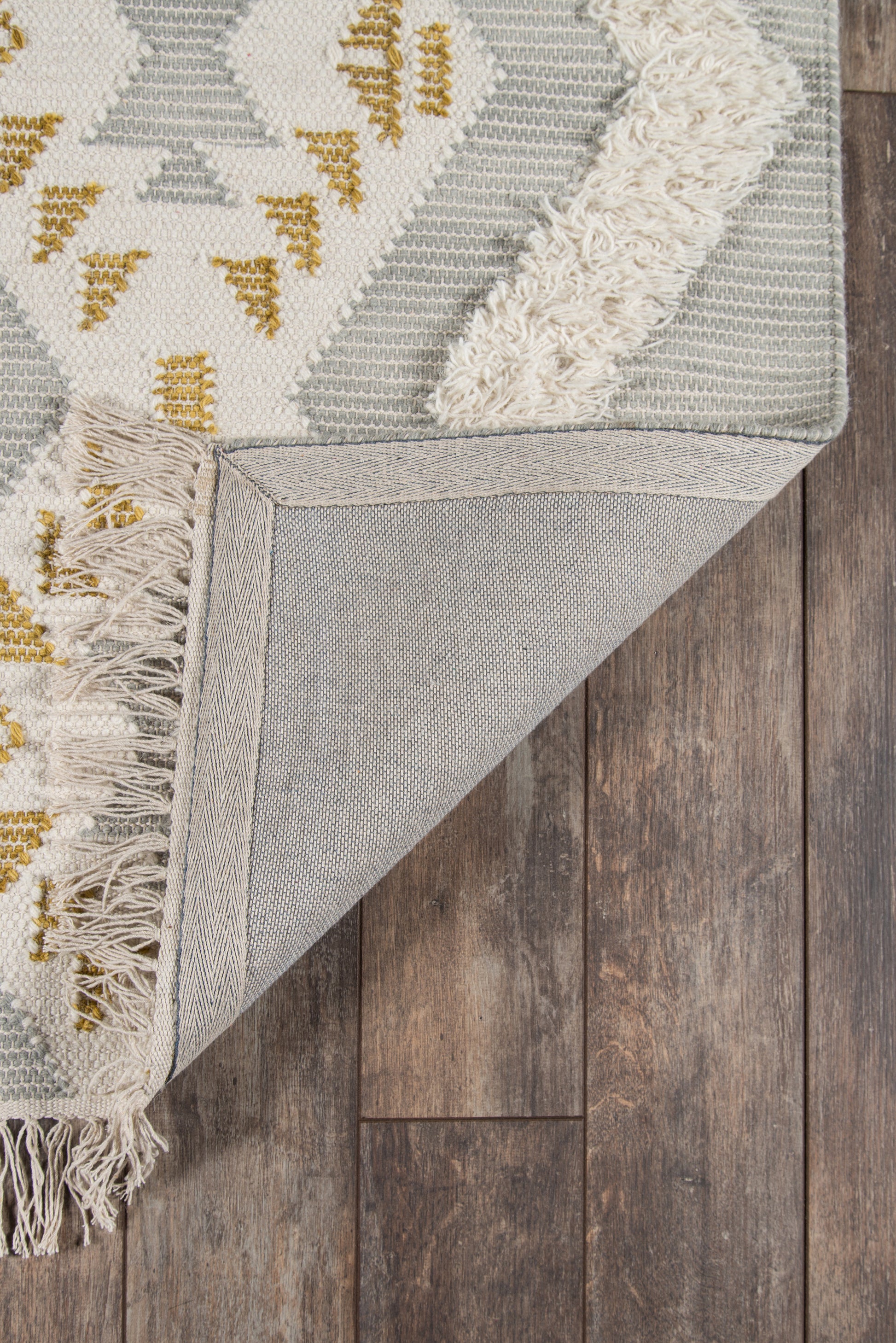 Ivory and gray geometric area rug with mustard accents, fringed edges, folded at corner to reveal woven underside on wood floor.