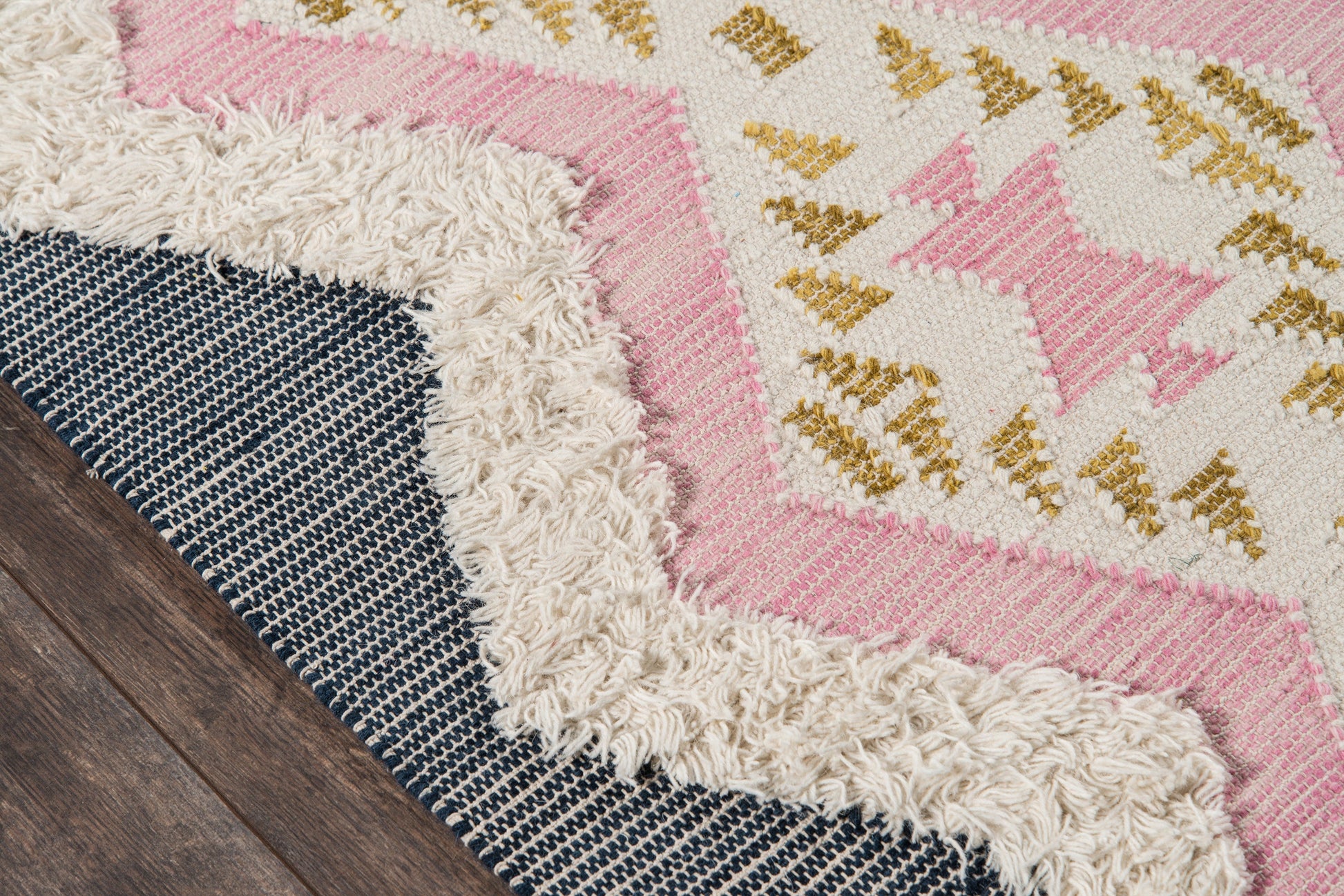 Boho area rug with pink and cream geometric pattern, gold accents, fluffy ivory fringe, layered on navy striped rug.