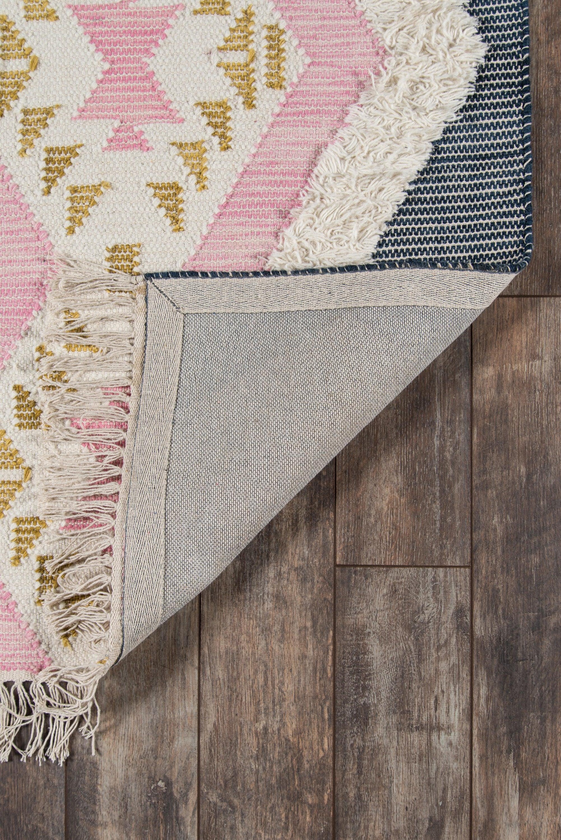Pink ivory and gold geometric woven rug with fringed edges; folded corner reveals gray underside and navy border on wood floor.