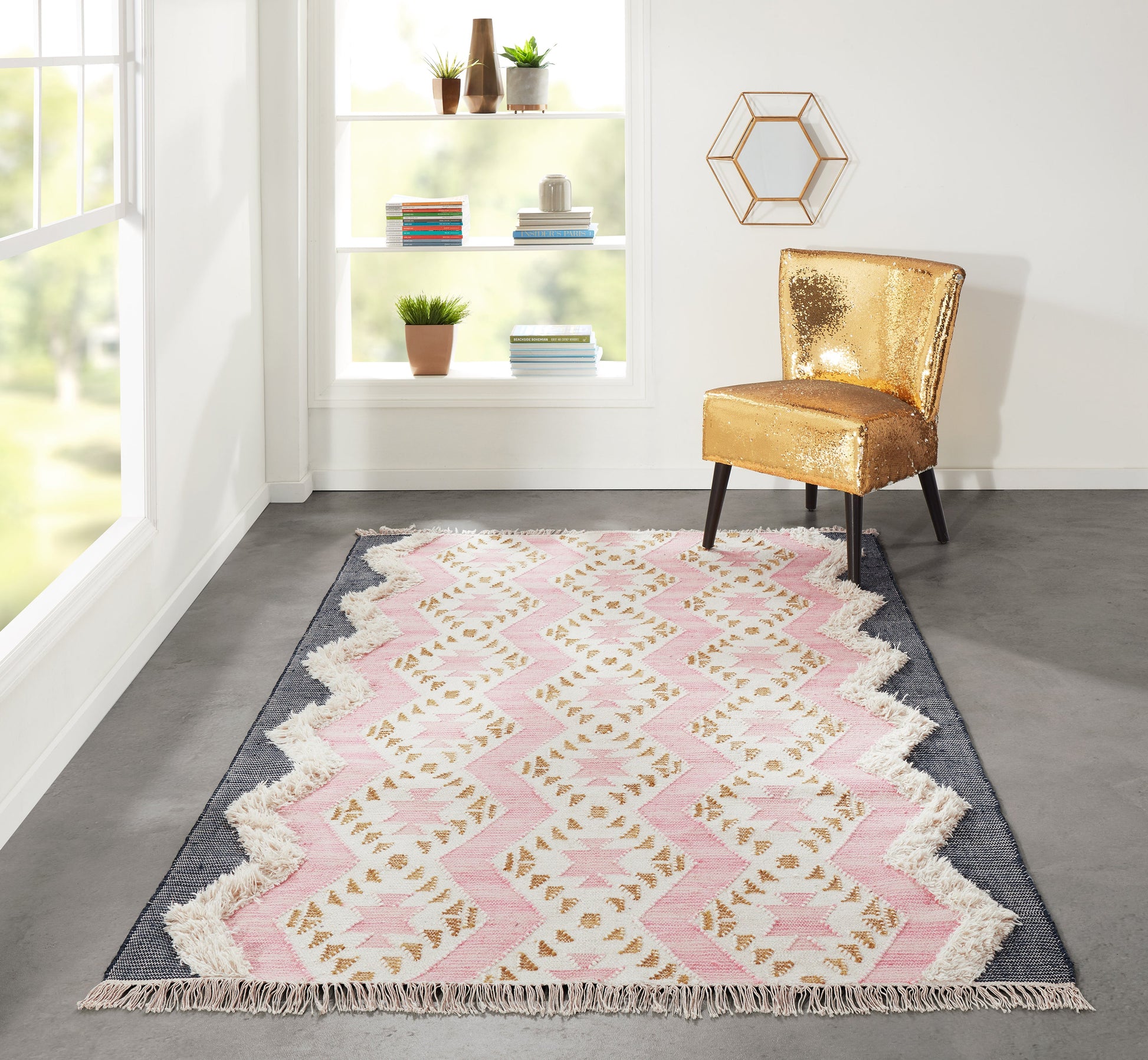 Pink geometric rug with white fringe on gray floor, gold sequin chair beside shelves of books and plants under hex wall art.