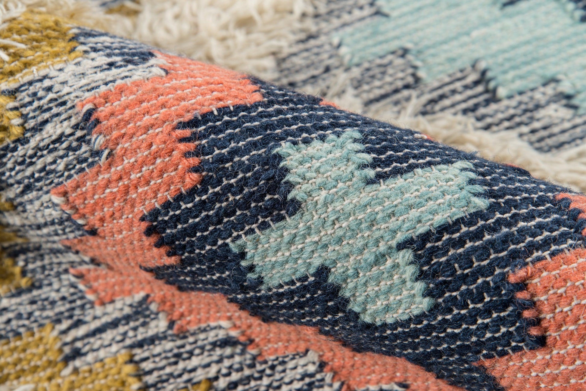 Close-up of a colorful woven rug with navy, coral, mint and mustard geometric diamonds, chunky yarns, fringed edge.
