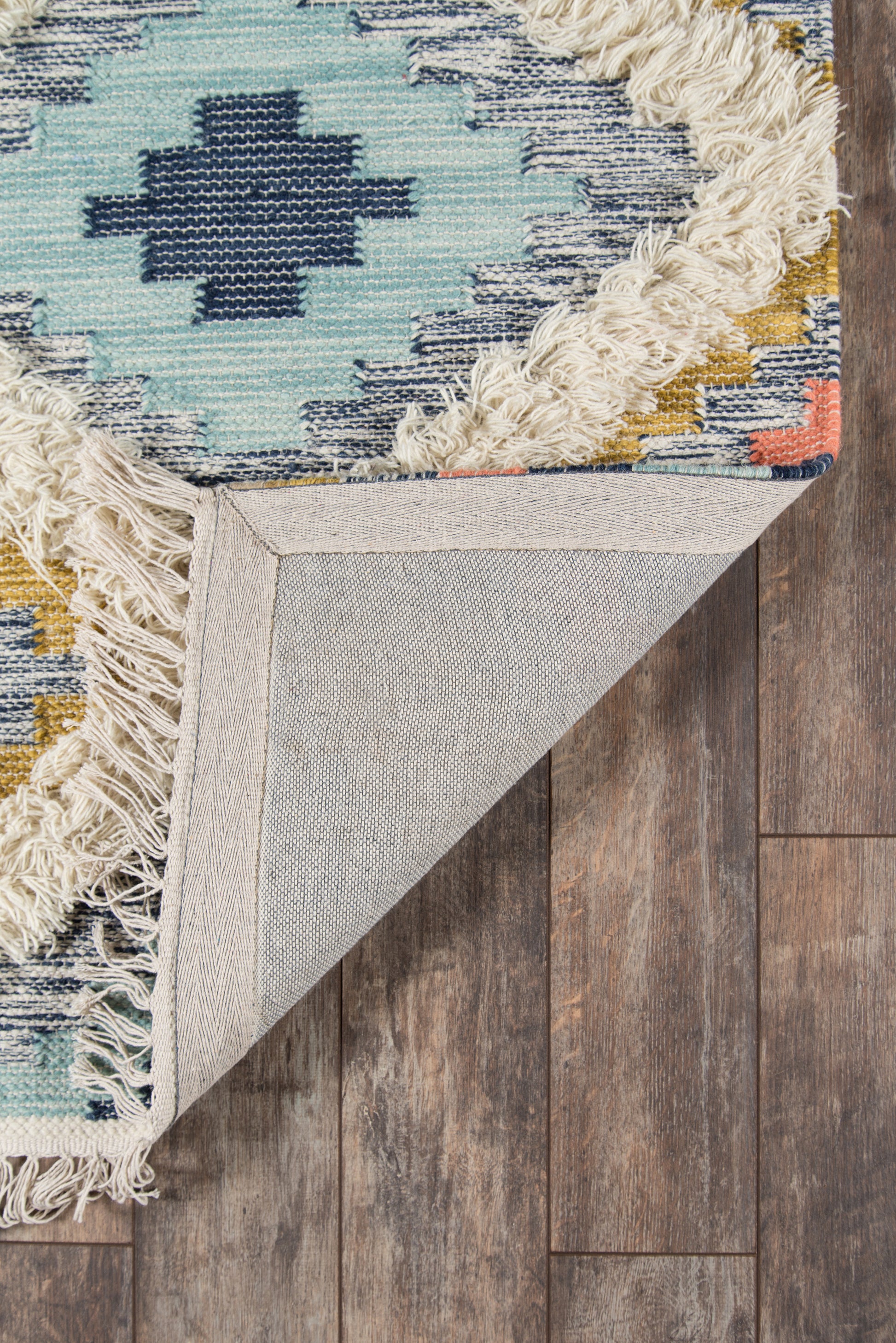 Boho geometric rug in turquoise and navy with cream fringe; woven textile, beige backing visible on wood floor.