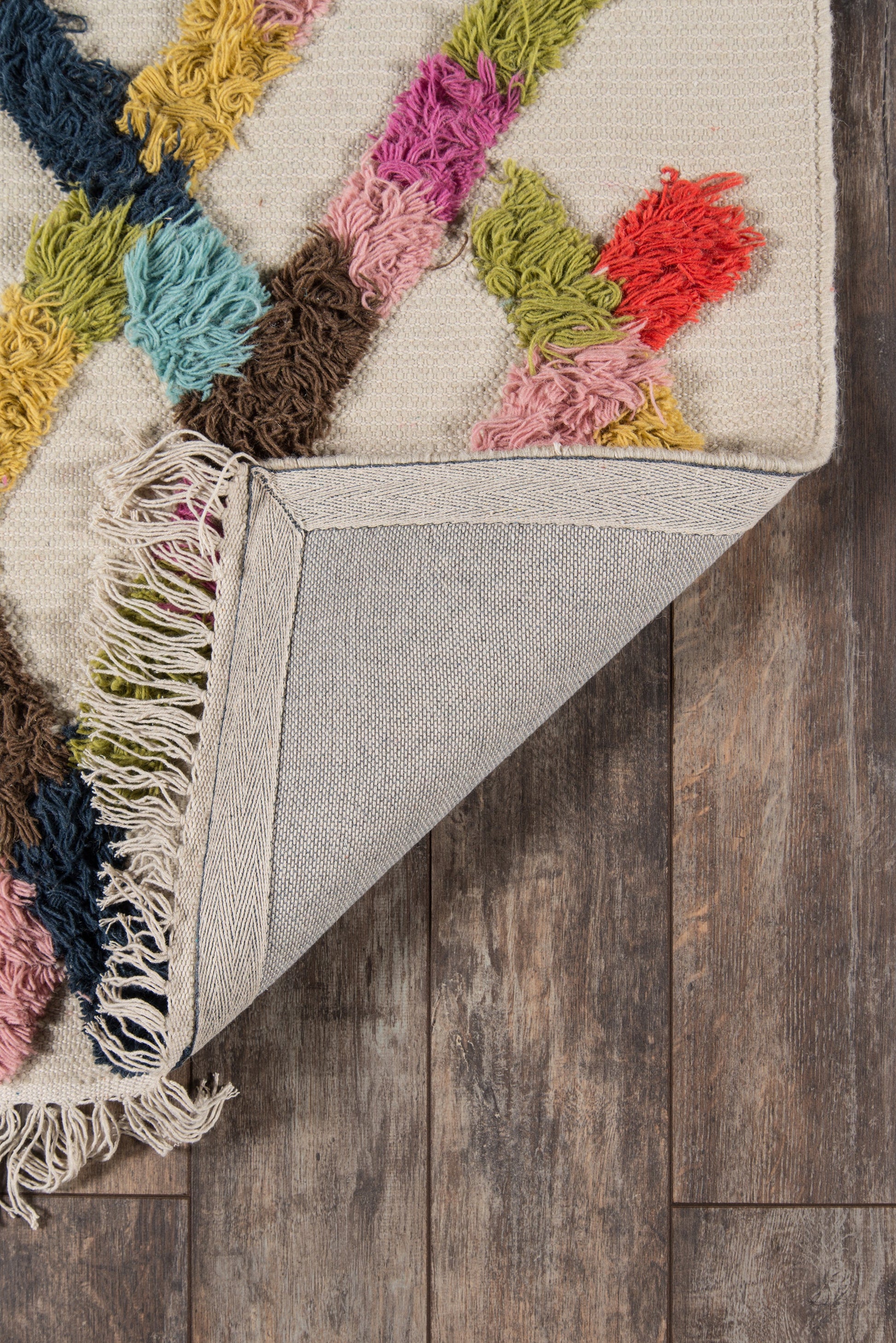 Beige tufted rug with multicolor chunky yarn tassels and fringe, folded corner showing light gray backing on wooden floor.