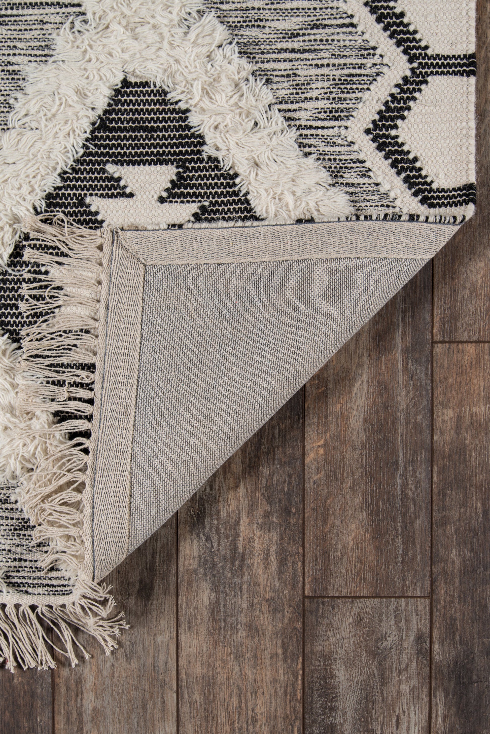 Black and cream geometric woven rug with fringe edges, folded to reveal beige underside on dark wood floor.