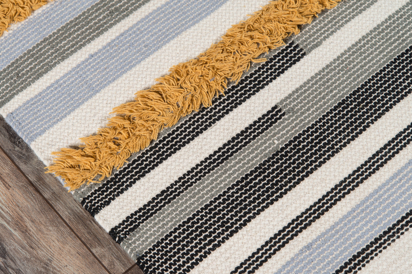Woven striped rug with cream, black, gray, and soft blue stripes; mustard fringe along edge, on wood floor.