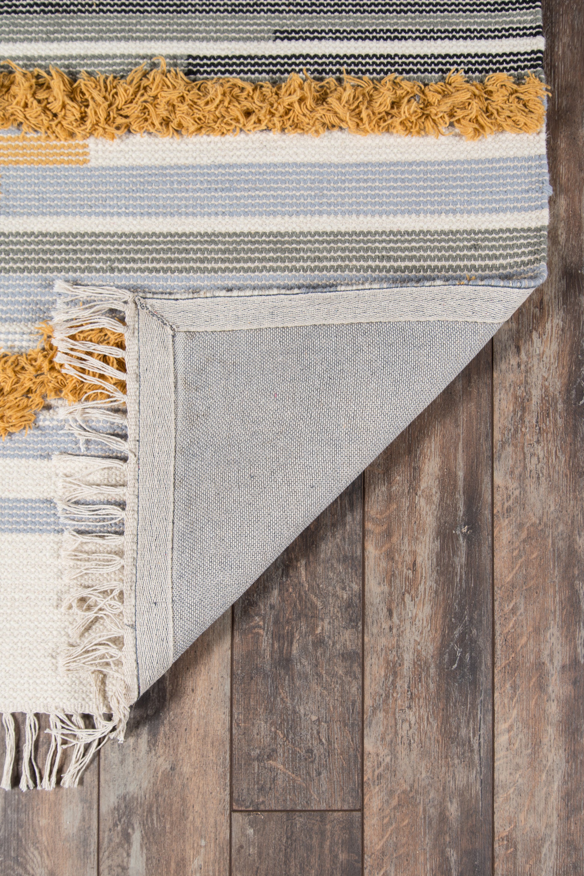 Striped woven rug with blue, cream and gray stripes, mustard fringe; folded corner reveals denim-like backing on wood floor.