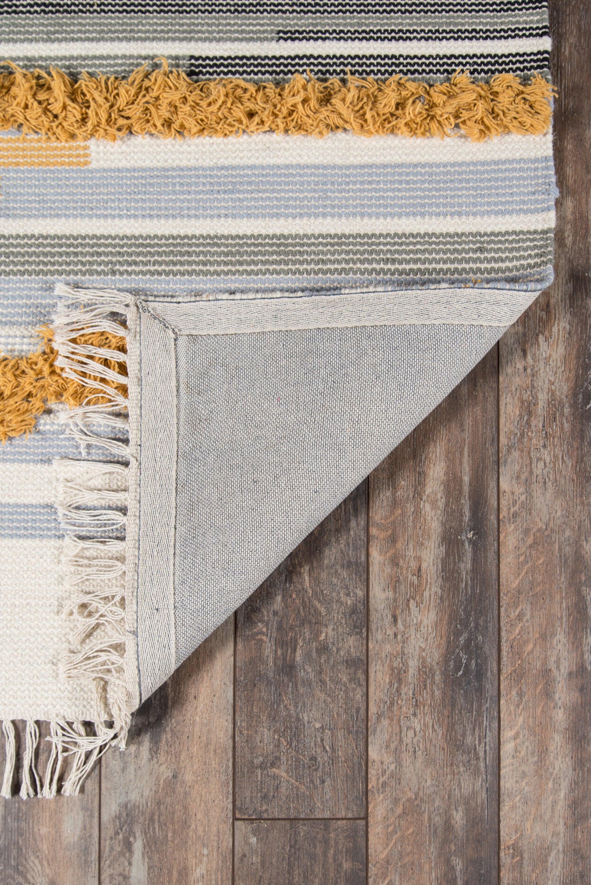 Blue, ivory and gray striped woven rug with mustard fringe, folded corner showing underside on wood floor.