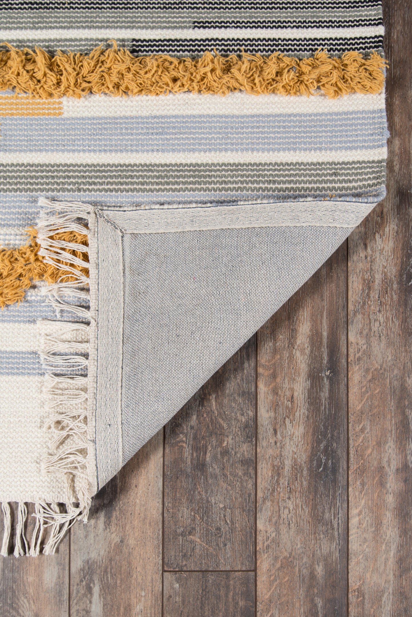 Blue, gray, white striped woven cotton rug with mustard fringe; corner folded to reveal denim underside on wood floor.