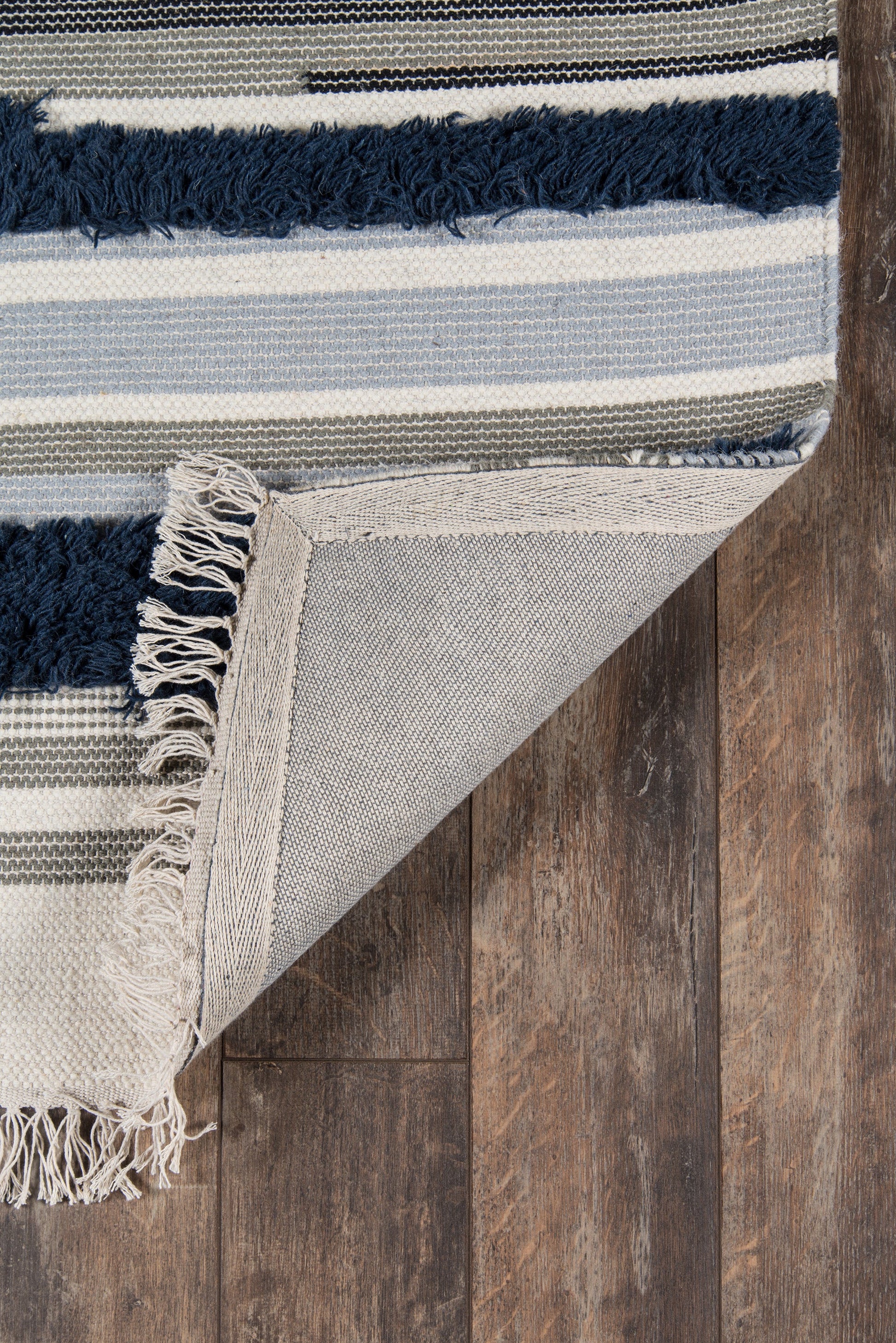 Striped woven rug with navy fringe, ivory, blue & gray stripes, folded corner on dark wood floor.
