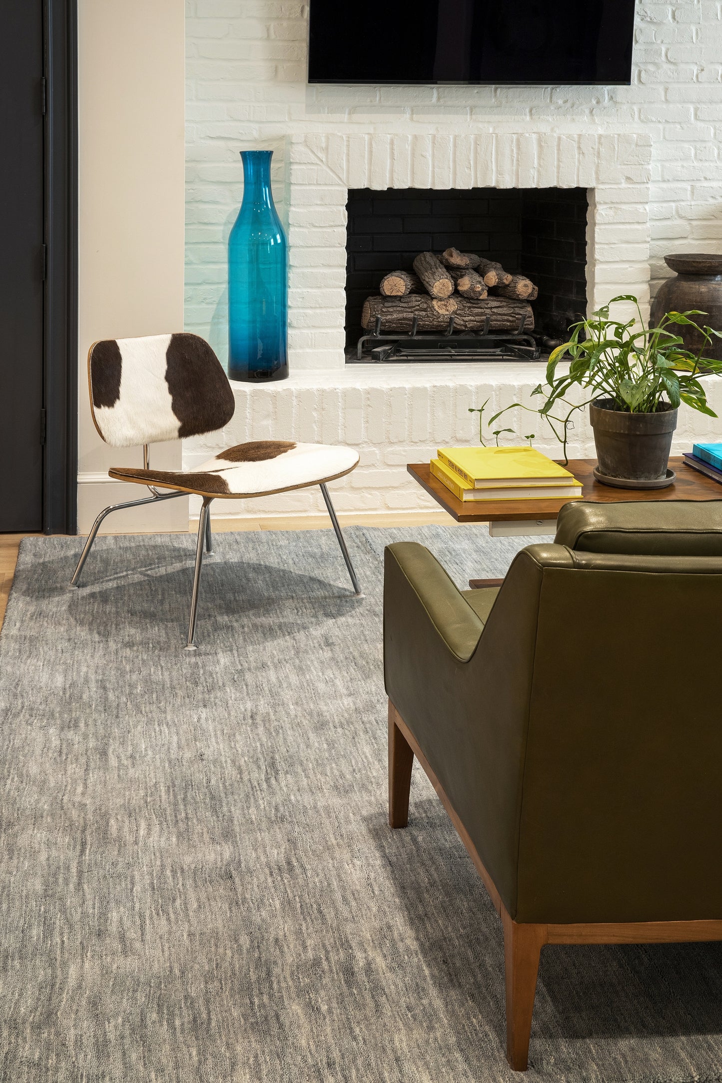 Mid-century living room with cowhide chair, olive leather sofa, turquoise vase, white brick fireplace and yellow books.
