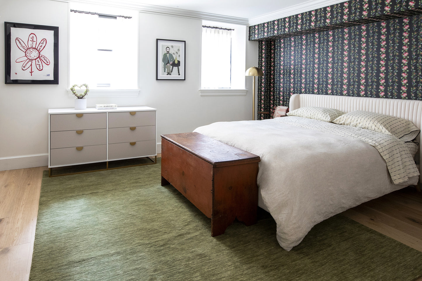 Bedroom with tufted white headboard, green rug, wooden bench, gray bedding, floral wallpaper, white dresser and framed art.