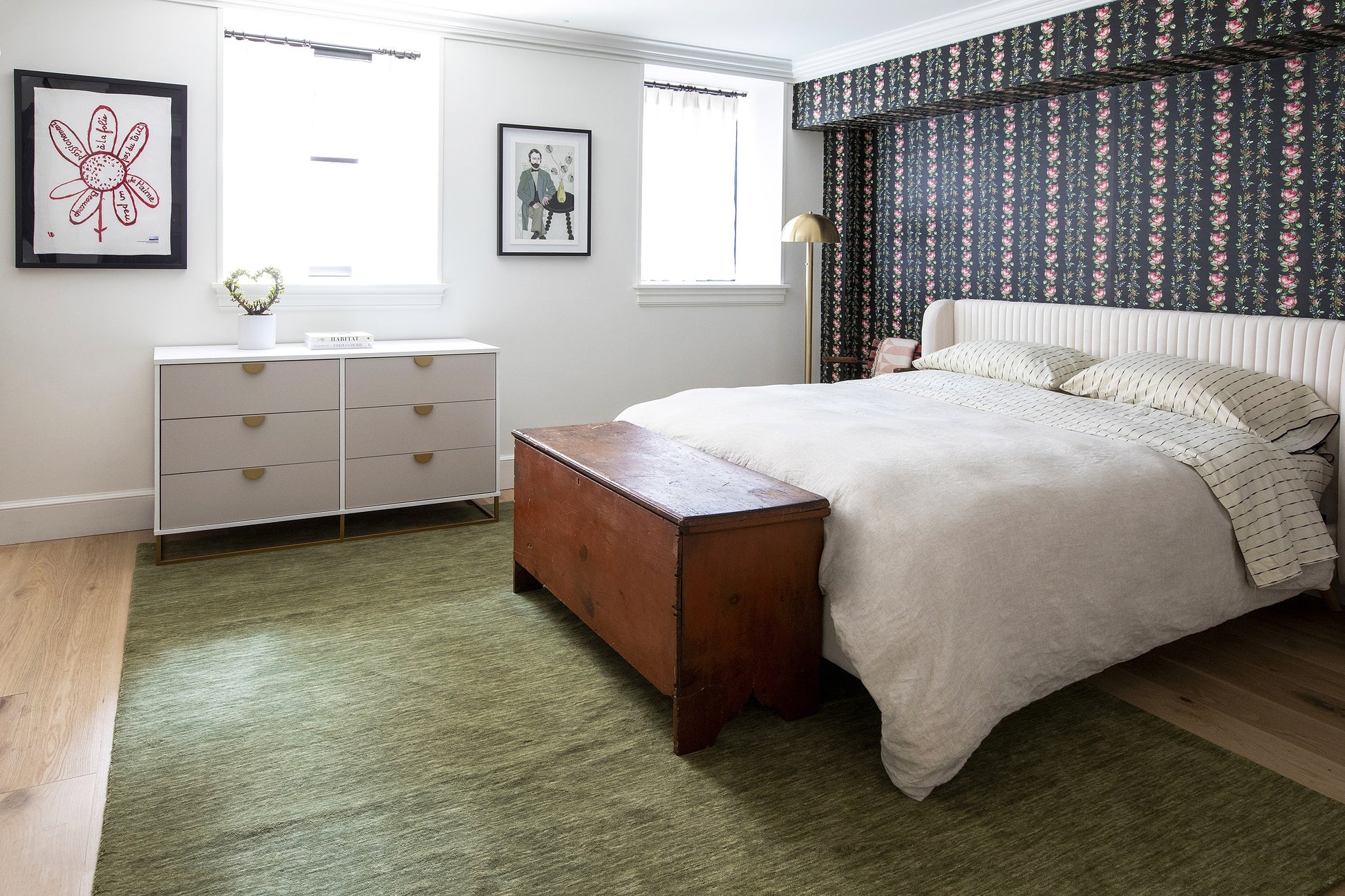 White bed frame with cream bedding, wood storage bench at foot, green rug, white dresser, framed art, and floral wallpaper.