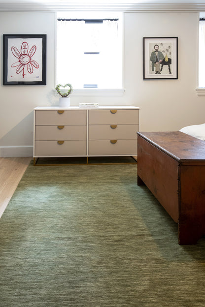 Bedroom scene with a beige 6-drawer dresser, wooden bed, green area rug, two framed artworks, and a small plant on the dresser.