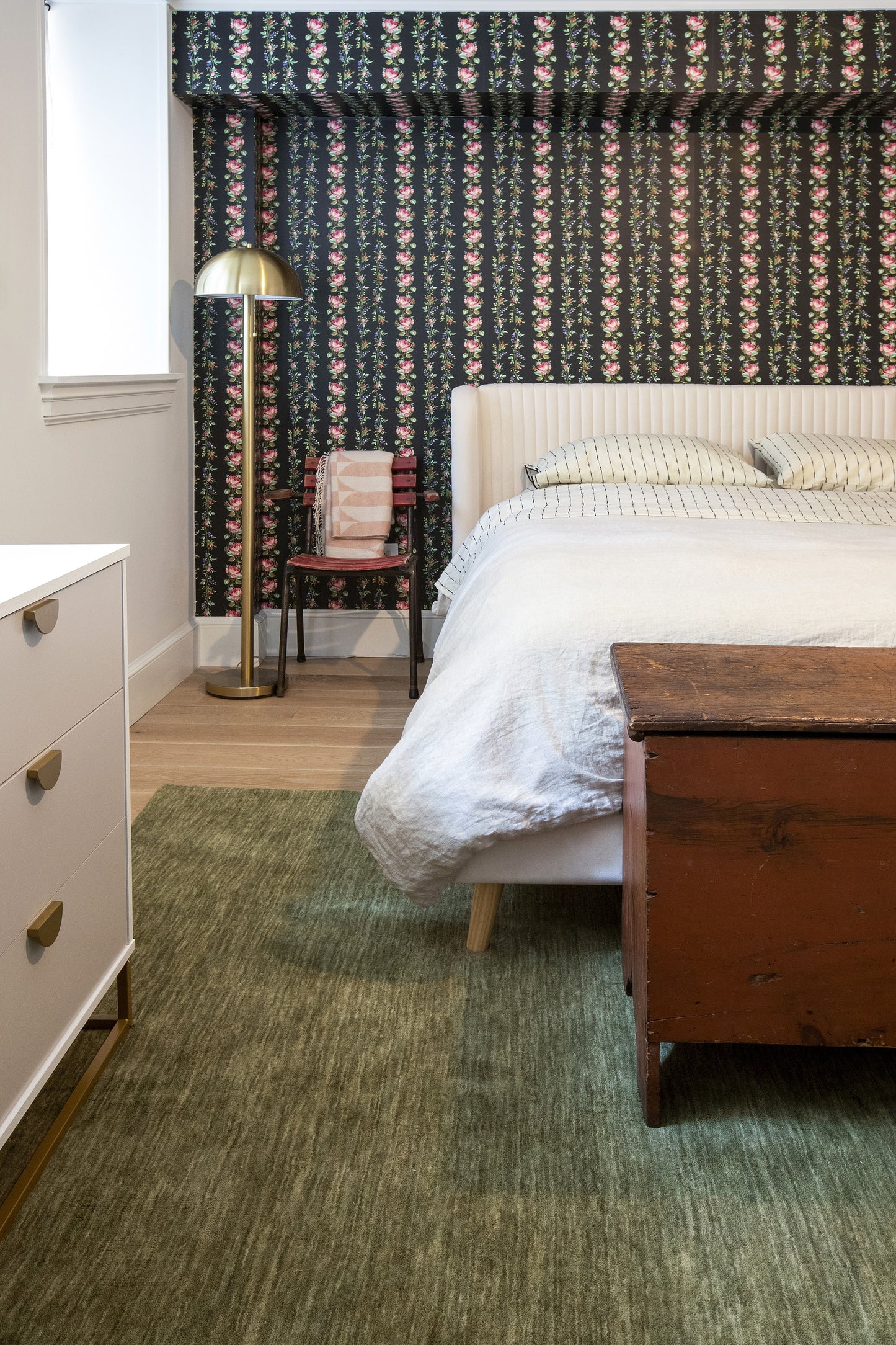 Bedroom scene with a green Iris 2' x 8' runner rug, white upholstered bed, wooden chest at foot of the bed, brass floor lamp, white dresser, and dark floral wallpaper.