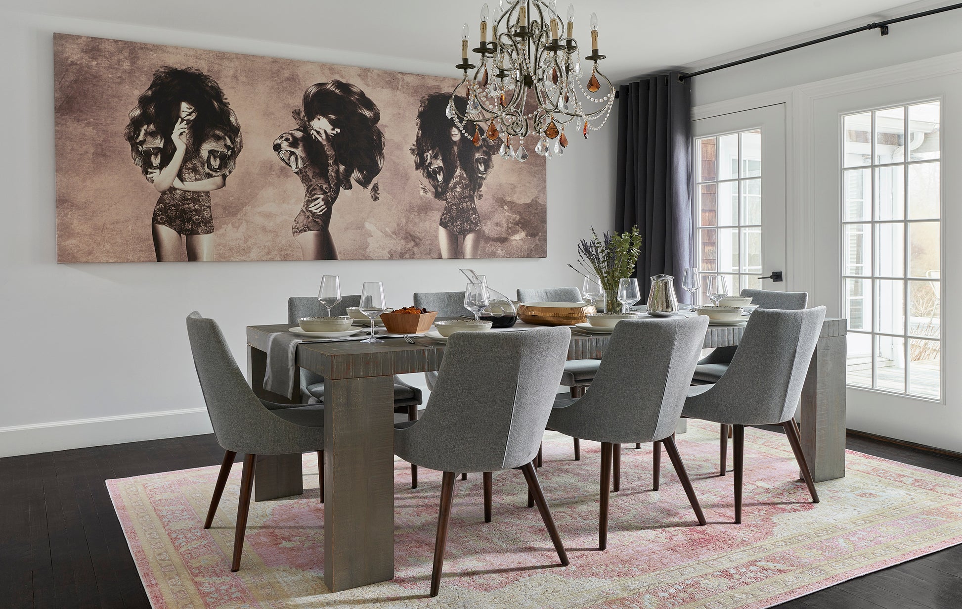 Contemporary dining room with a long gray wood table, gray upholstered chairs, chandelier, large wall art, and pink rug.