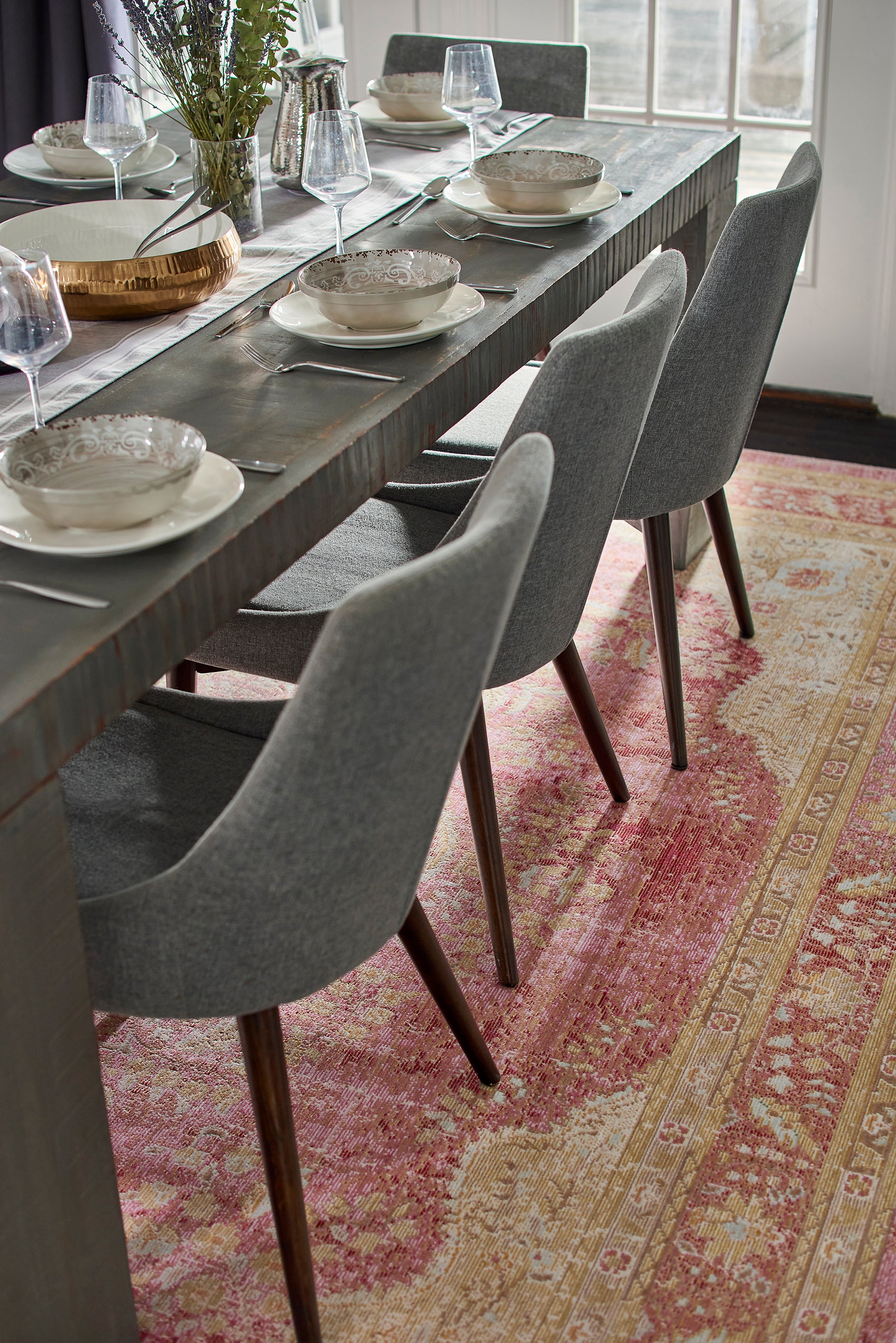 Pink patterned rug under a long dark wooden dining table with gray upholstered chairs, bowls, glassware & cutlery.