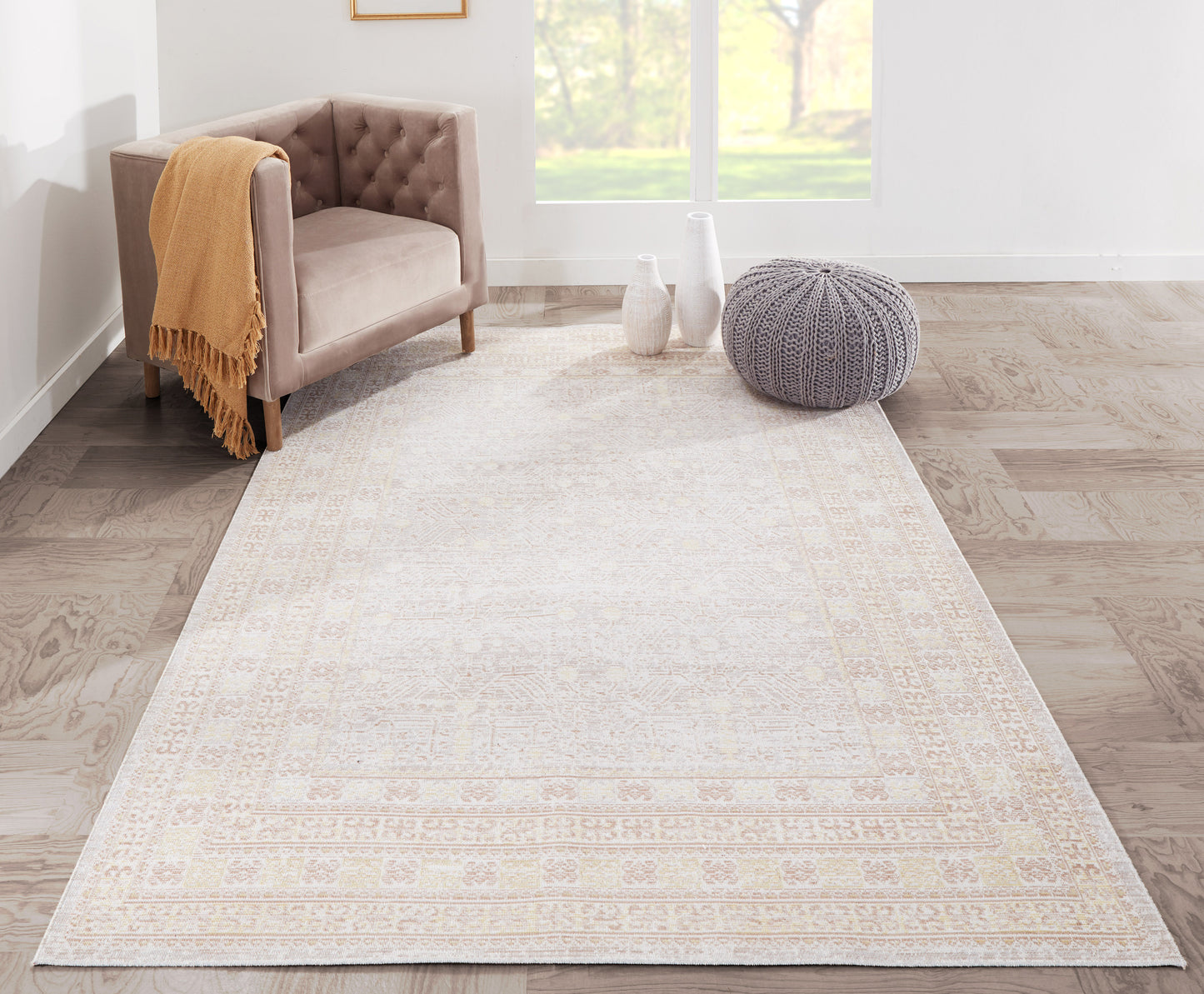 Beige-gray area rug with a subtle border in a bright living room, tan tufted chair and gray knit pouf.