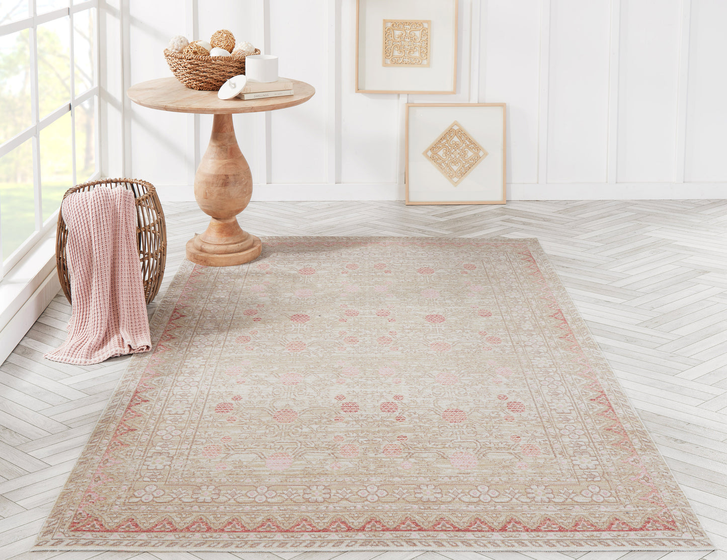 Beige oriental rug with pink floral motif and border, in bright room with round wooden table, wicker basket and pink throw.