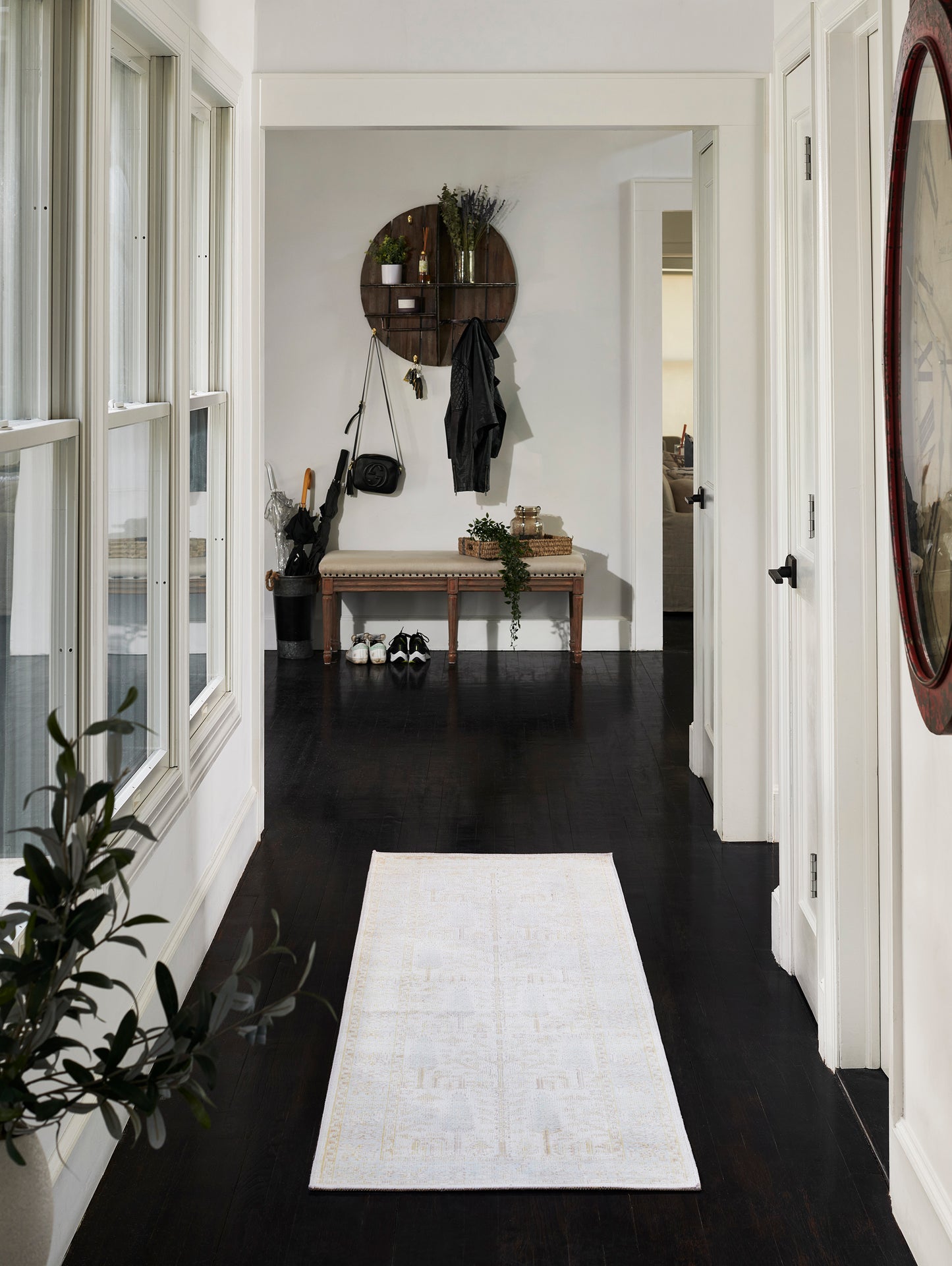 White hallway with dark wood floors, pale patterned runner, wooden bench, round wall shelf with coats and plants, red mirror.