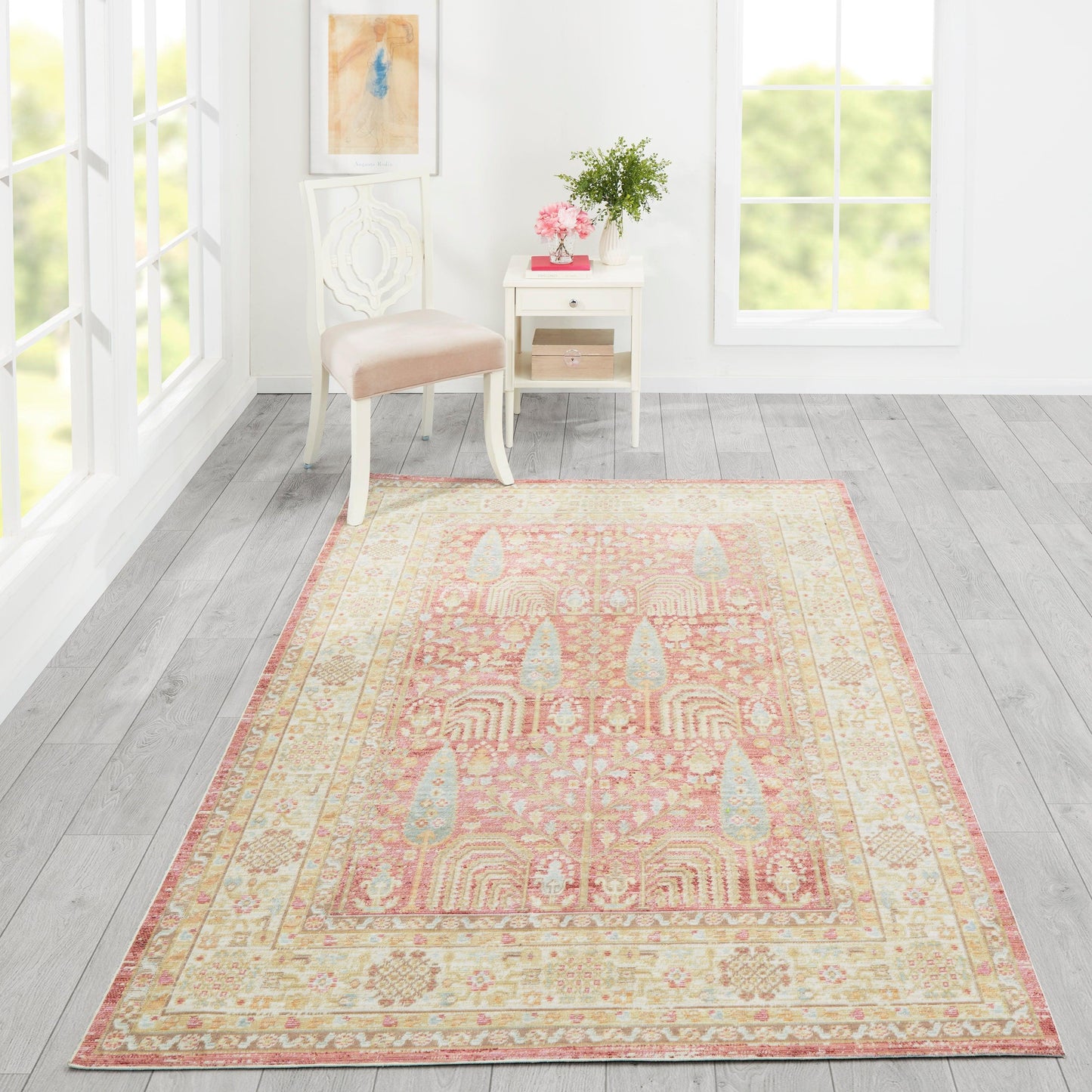 Soft peach-pink oriental rug with cream border on light gray wood floor, beside a white chair and table with pink flowers.