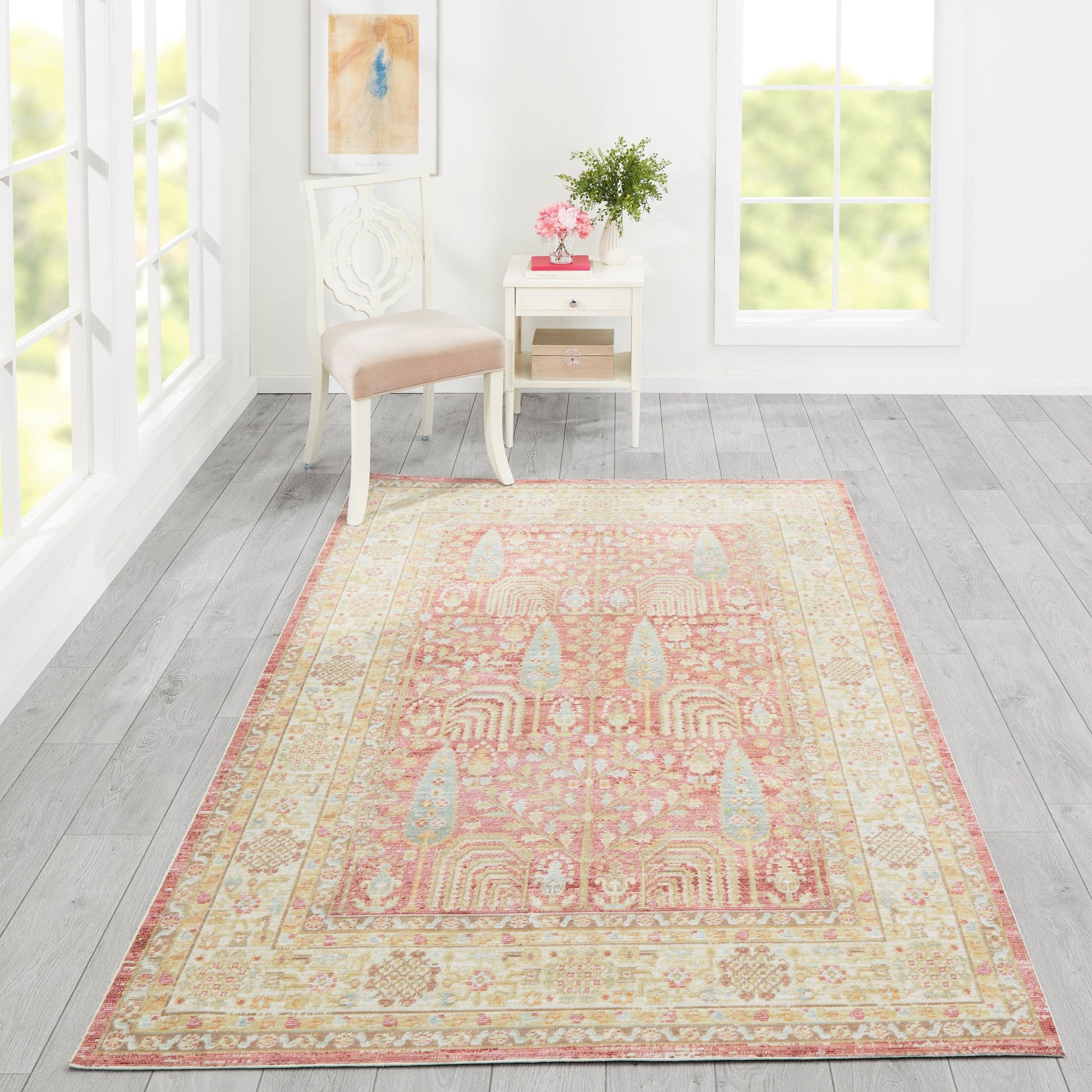 Soft peach-pink oriental rug with cream border on light gray wood floor, beside a white chair and table with pink flowers.