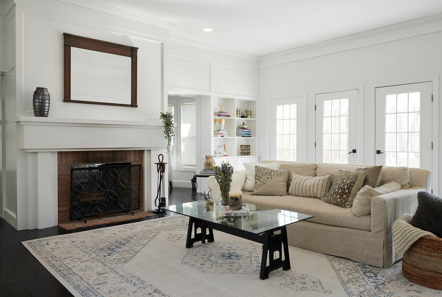 Bright living room with beige sectional, glass coffee table, white fireplace, built-in shelves, and a blue-patterned area rug.