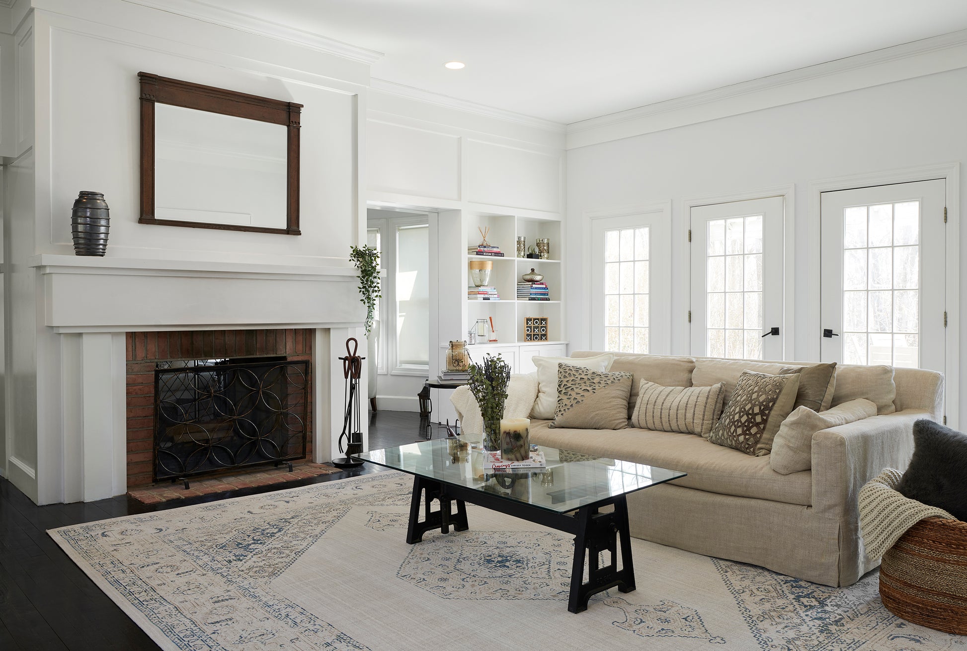 Bright living room with beige sectional, glass coffee table, white fireplace, built-in shelves, and a blue-patterned area rug.