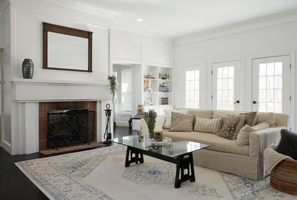 Bright living room with beige sectional, glass coffee table, white fireplace, built-in shelves, and a blue-patterned area rug.