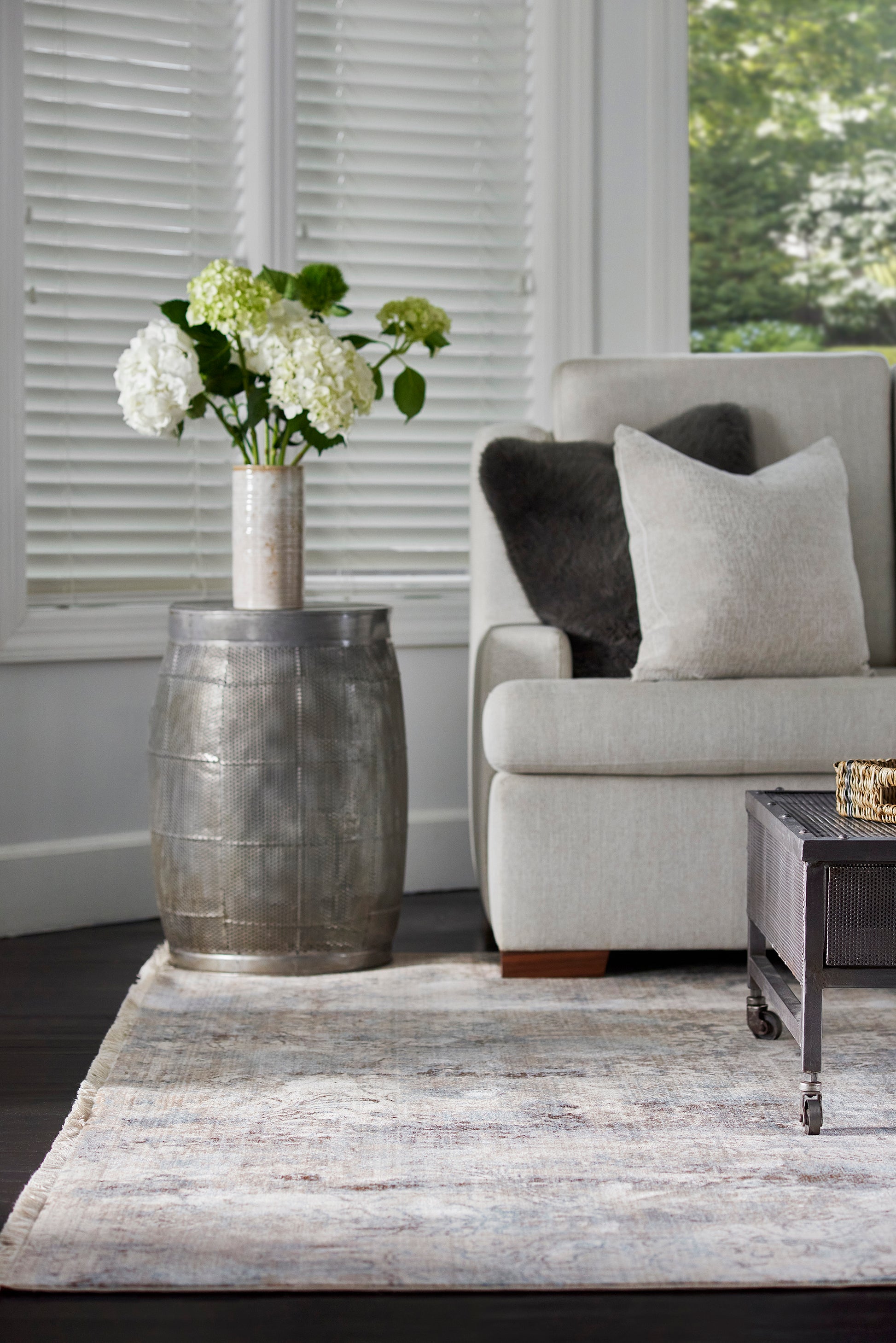 Industrial metal barrel side table with a tall metallic vase of white hydrangeas beside a light gray sofa.