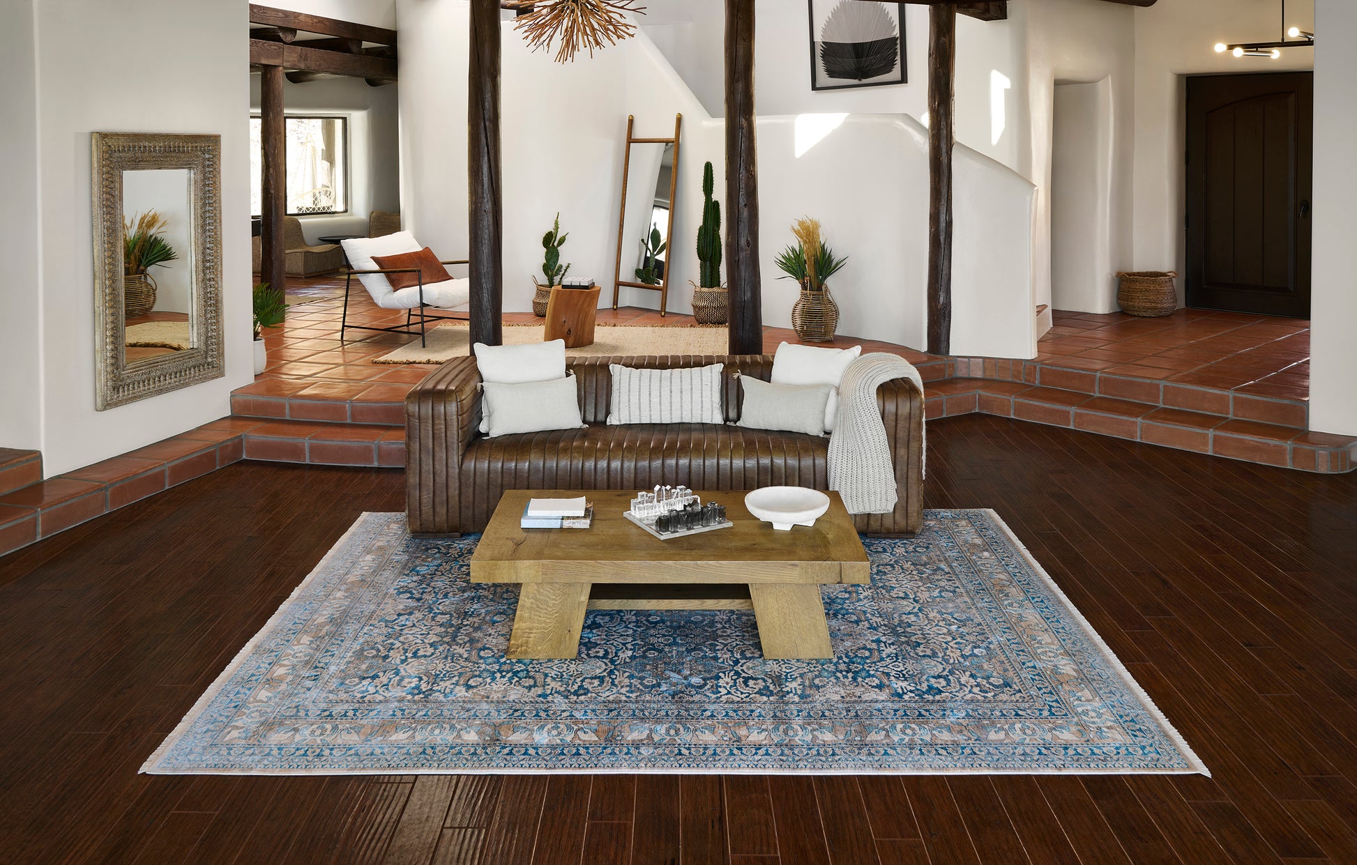 Wicker sofa with white cushions on a blue patterned area rug, wooden coffee table, dark hardwood floor, beams.