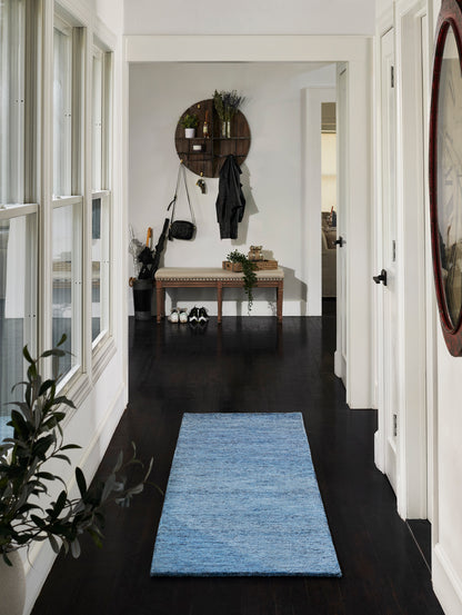Blue woven entryway runner rug on dark hardwood floor, in a bright hallway with white walls and a bench plus coat rack.