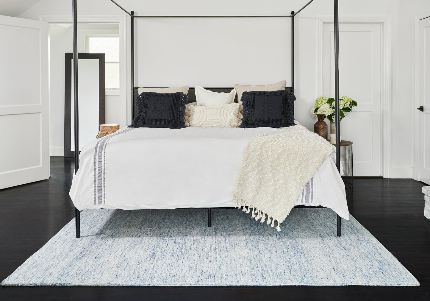 Black metal four-poster bed with white bedding, black and cream pillows, cream knit throw; light blue rug on dark floor.