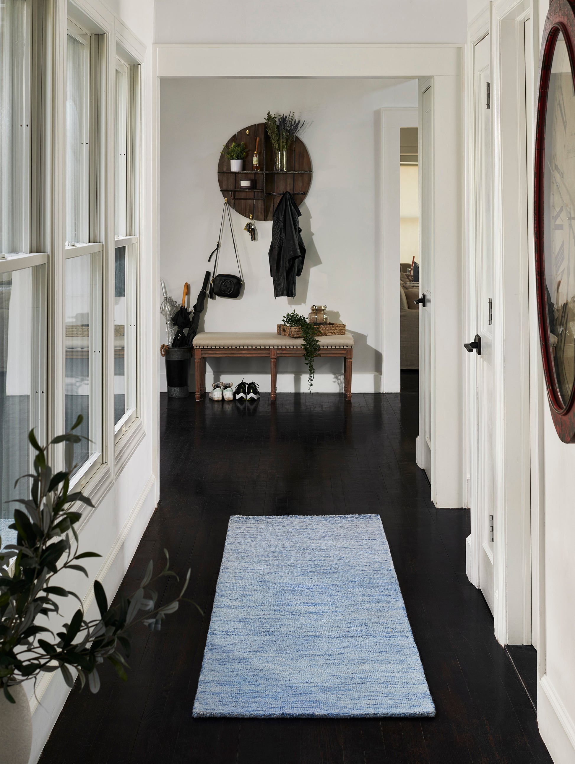 Bright entryway with dark wood floors and a light blue 8' x 10' rug, wooden bench, round wall shelf with hooks, coat and bag, and plants.