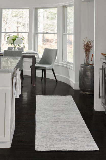 Light gray woven rug runner on dark wood floor beside a white kitchen with gray upholstered dining chairs by bay windows.