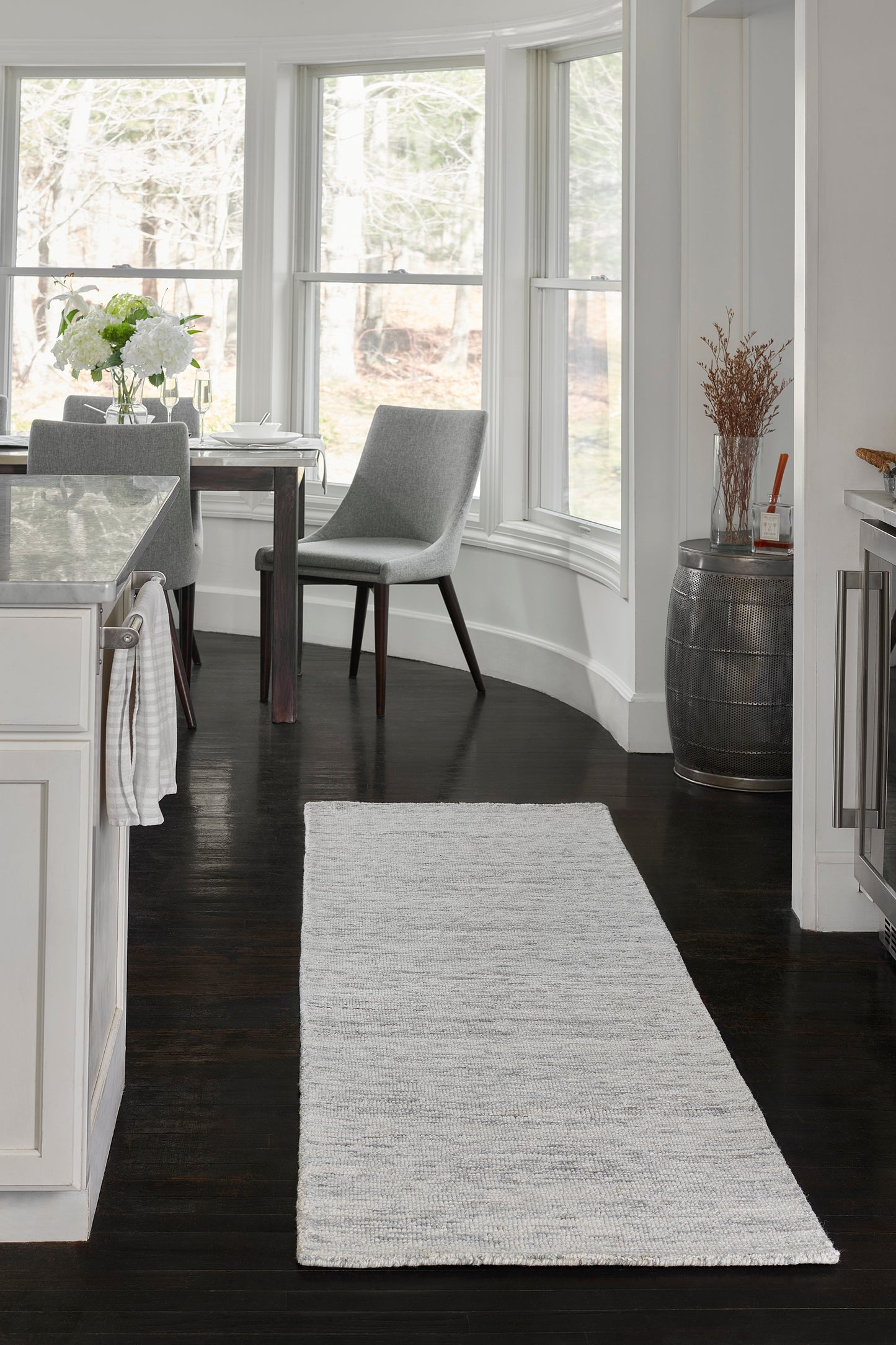 Soft light gray rectangular runner rug on dark wood floor in a white kitchen with glass dining table and gray chairs.