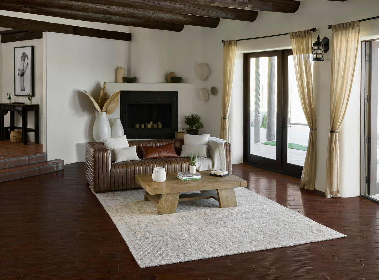 Cozy living room with brown leather sofa, beige rug, wood beam ceiling, fireplace, rustic coffee table, and cream curtains.