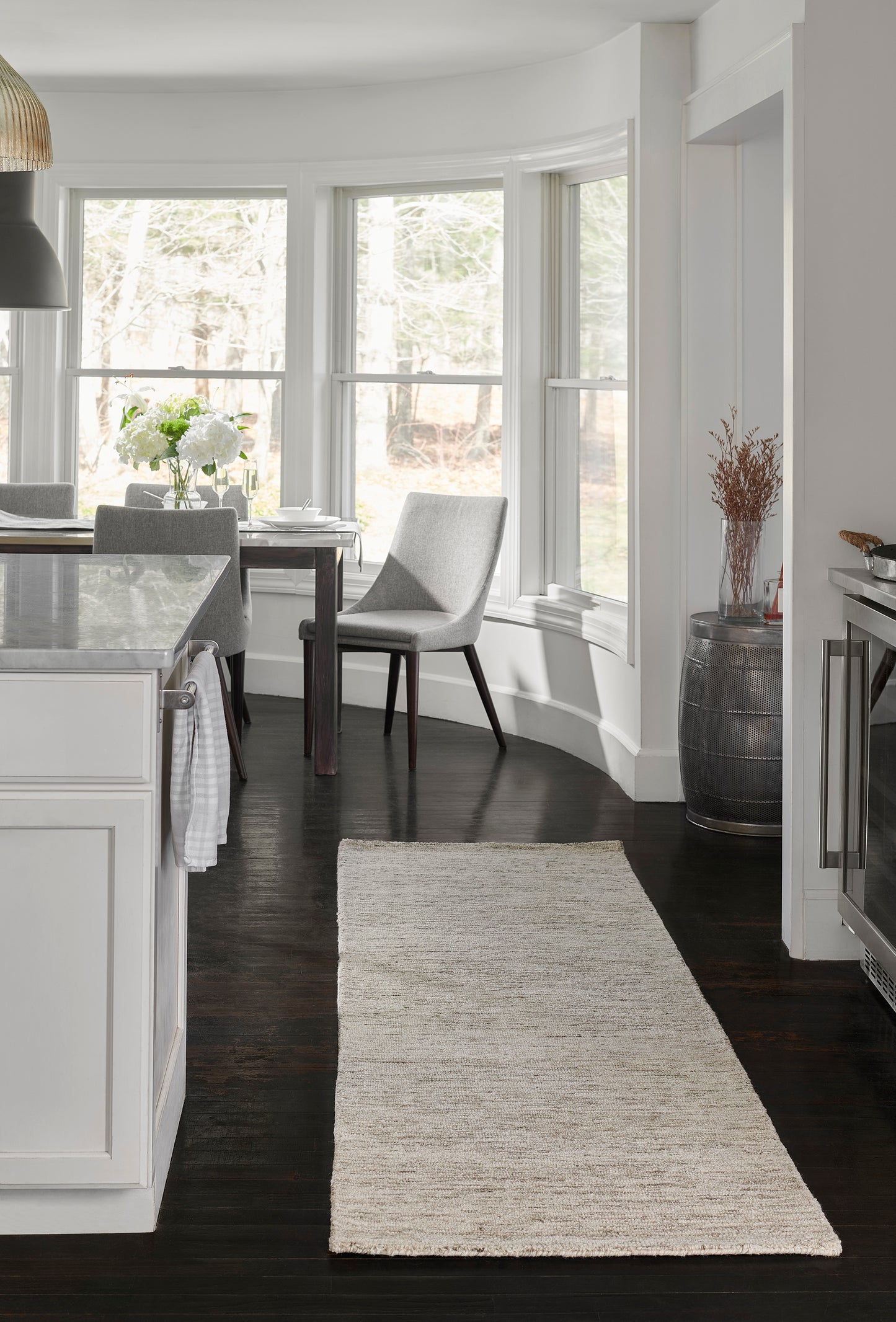 Neutral kitchen and dining area with white cabinets, marble island, dark wood floor, beige 9' x 12' area rug, gray chair by windows.