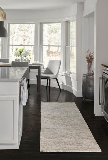 Bright white kitchen with dark wood floors, bay windows & a natural beige runner rug; gray dining chair near a marble island.
