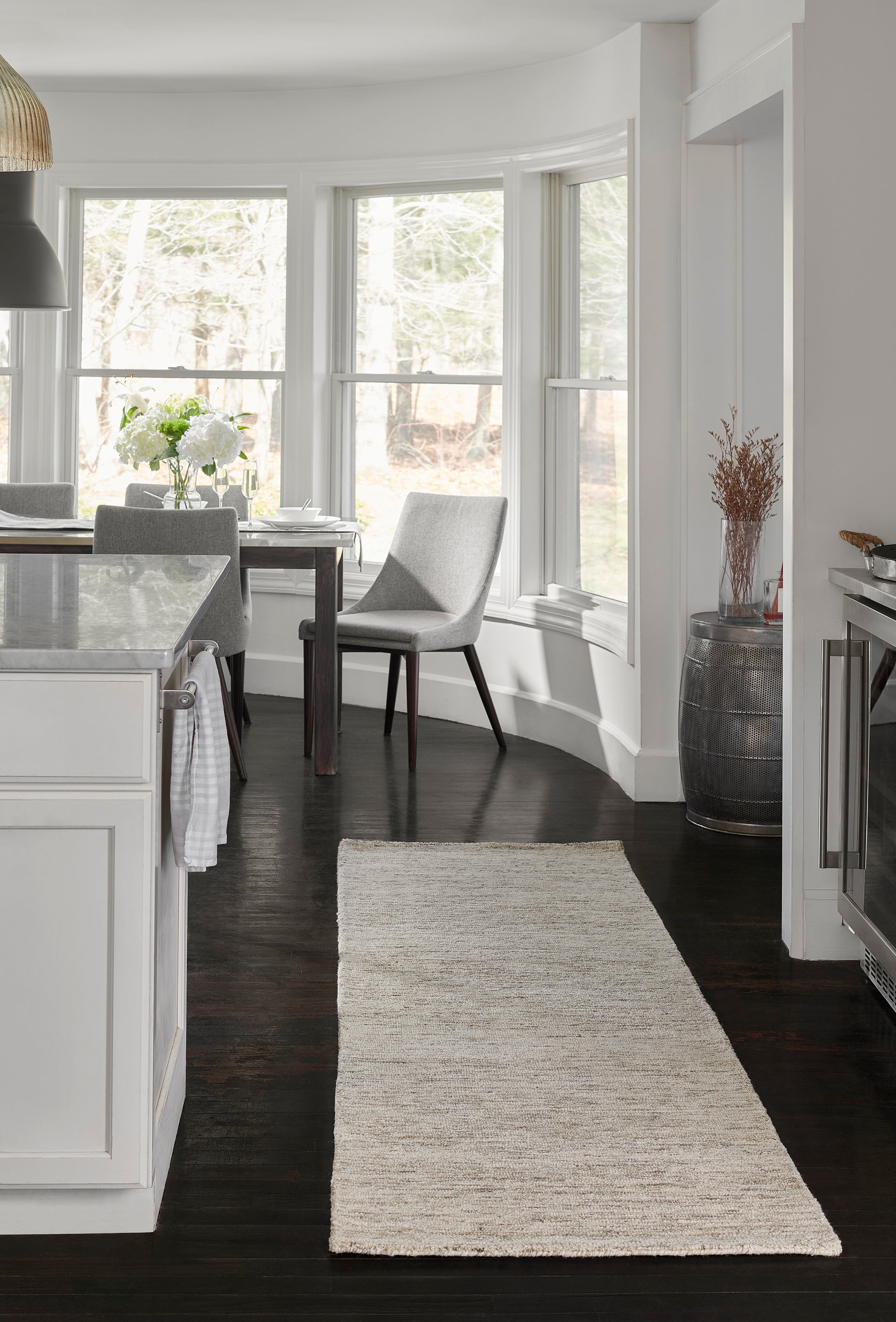 Bright modern kitchen with white cabinets, dark wood floors, beige runner rug, island & gray dining chairs.