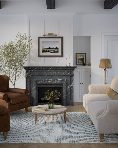 Cozy living room with dark marble fireplace, white paneling, beige sofa, brown leather chair, round coffee table, blue rug, plants.
