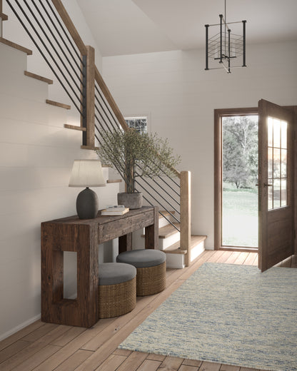 Rustic-modern entryway with wood console, wicker stools, potted olive tree, blue-gray runner rug, metal cable railing, open door.