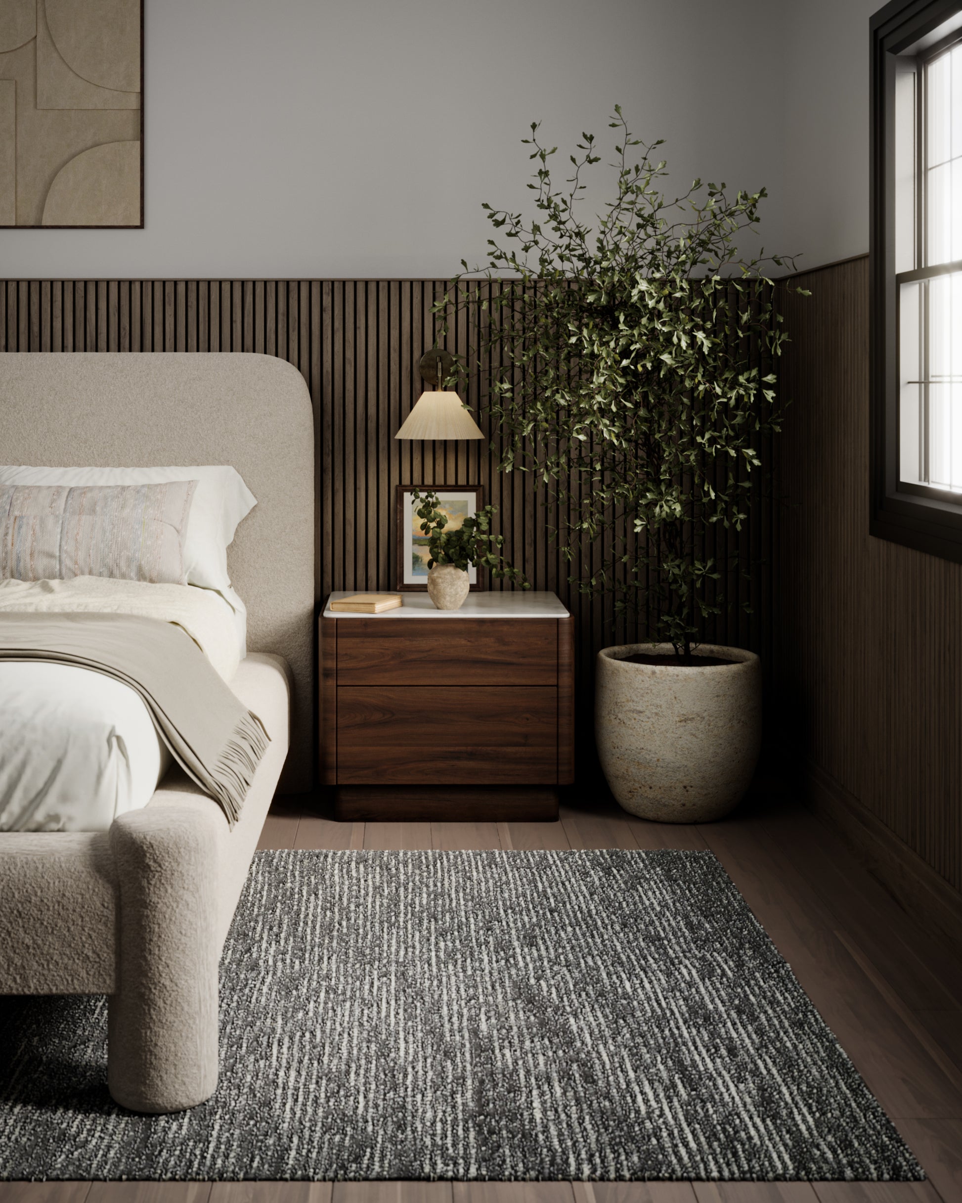 Beige upholstered bed with curved headboard, wooden nightstand, lamp, tall potted tree, and gray textured rug in a calm bedroom.