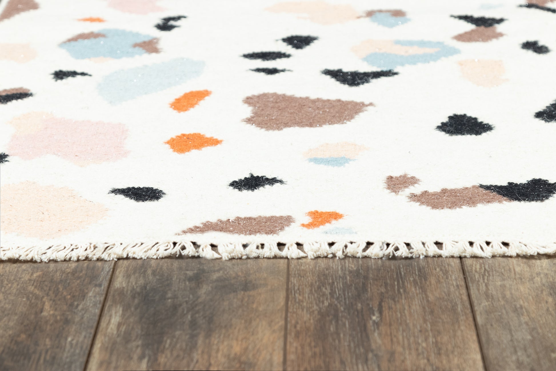 White rug with abstract multicolor spots (blue, pink, orange, beige, brown, black) and fringed edge on wooden floor.