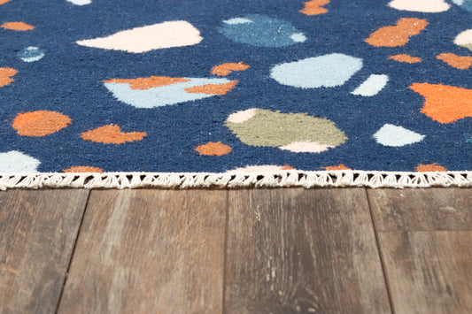 Blue rug with multicolor abstract shapes (orange, white, teal, olive) and fringe along a wooden floor.