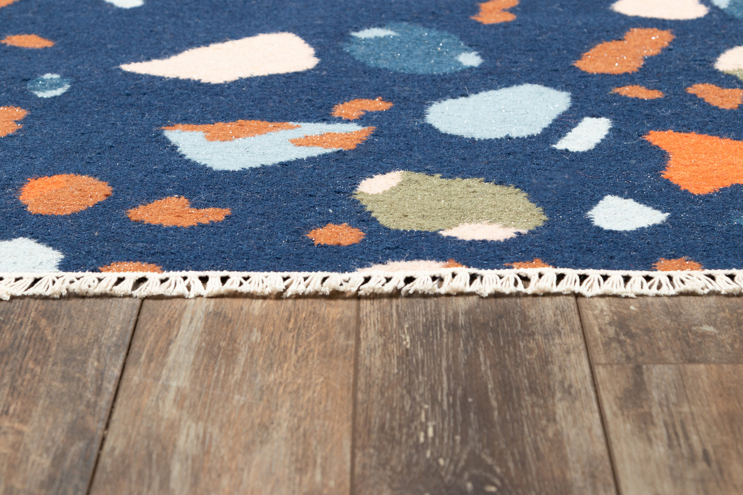 Navy blue abstract-pattern rug with orange, light-blue, white and beige blobs, fringe edge on a wooden floor.
