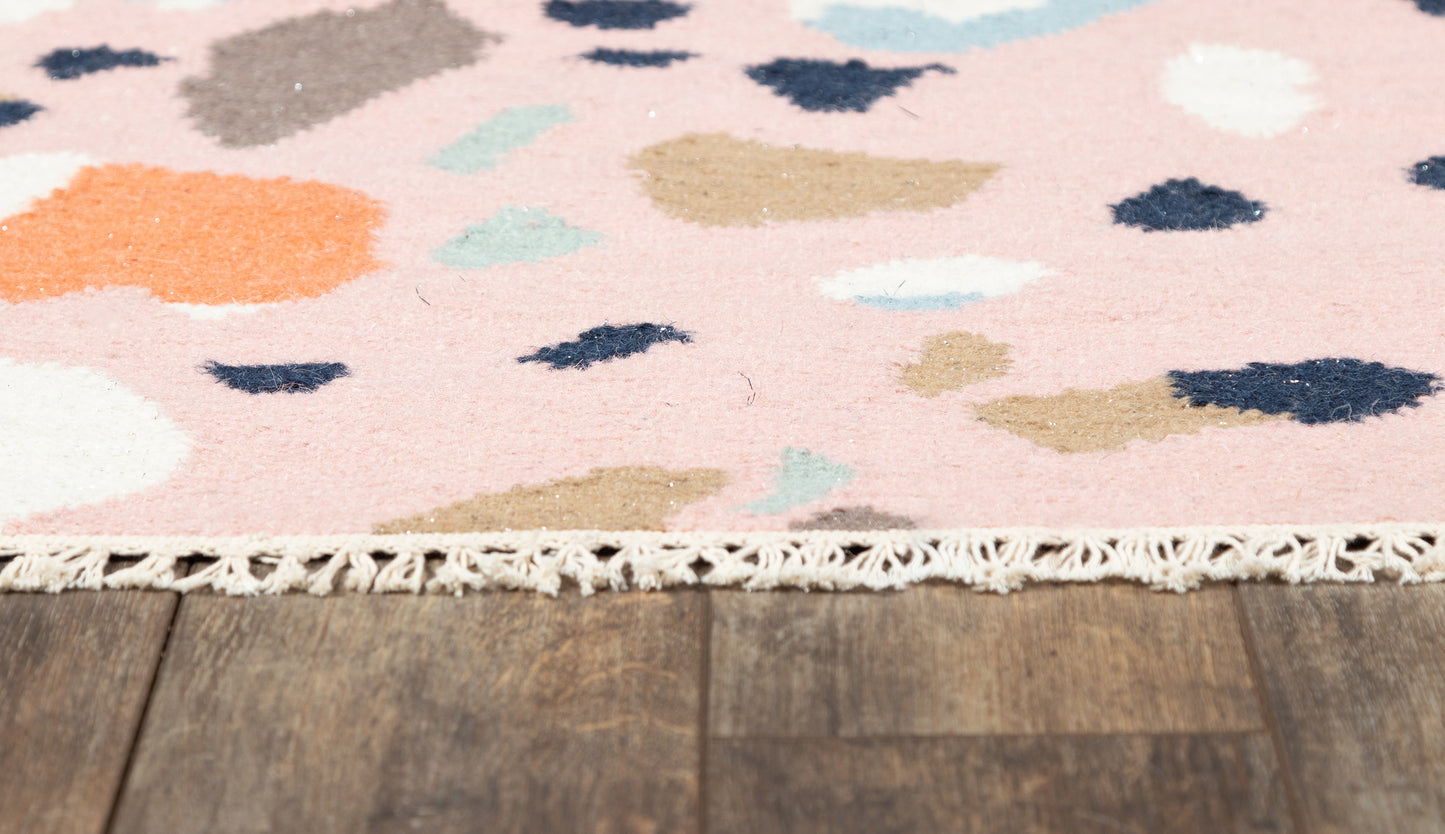 Pink abstract-pattern rug with multicolor blotches and a white fringed edge on a wooden floor.