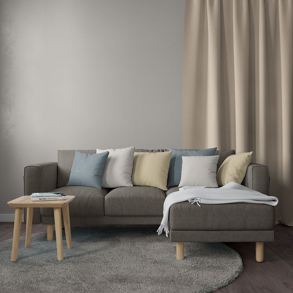 Latte solid fabric in a living room scene; L-shaped gray-brown sofa with pastel cushions, round light wood coffee table, gray rug, beige curtains.