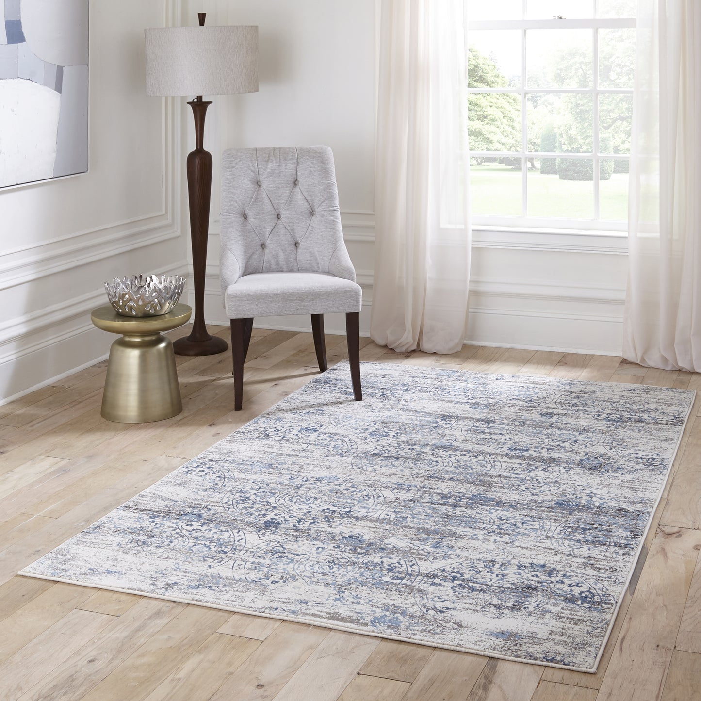 Light gray tufted chair with dark wood legs beside a brass side table and floor lamp on a blue-gray patterned area rug.