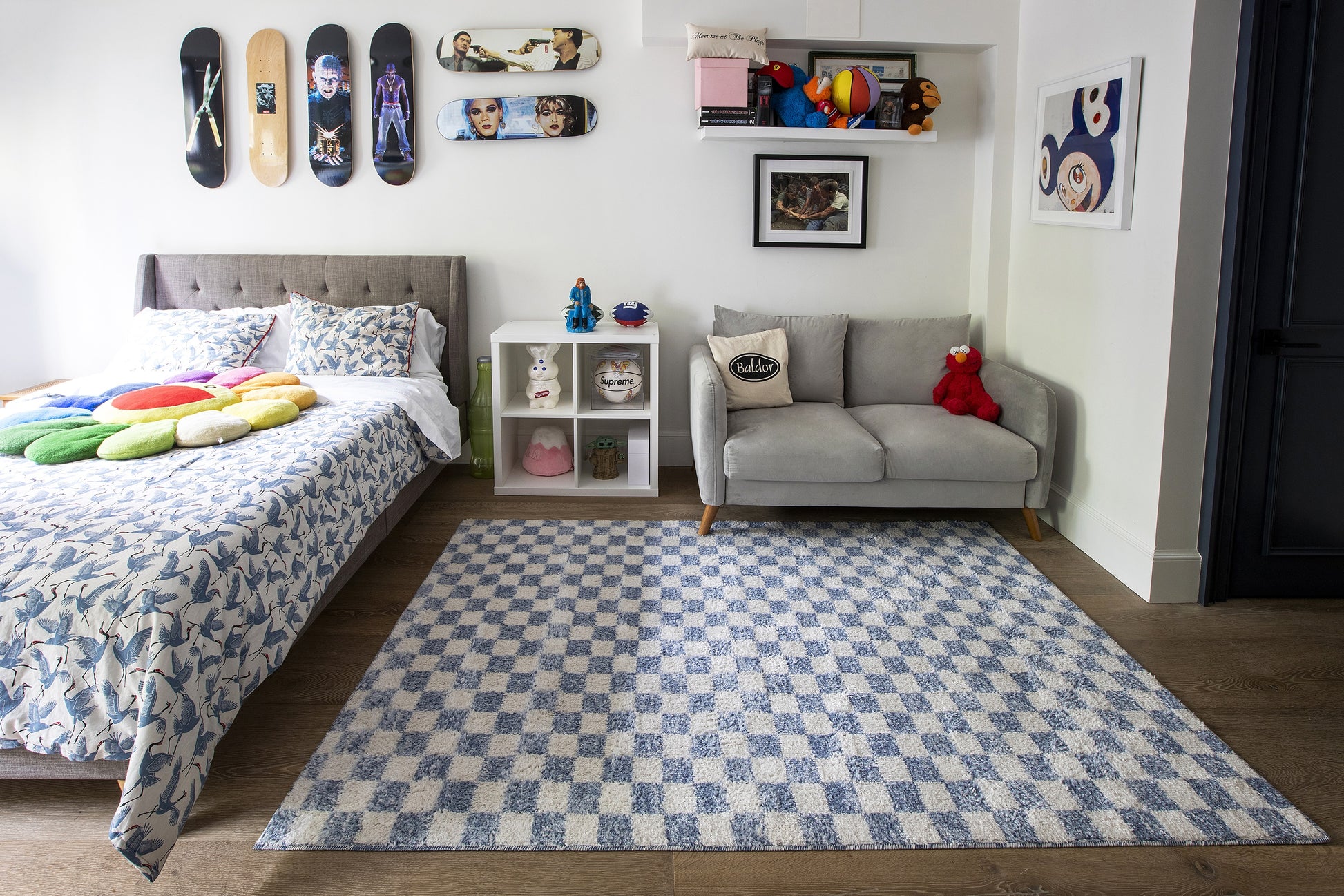 Kids’ bedroom featuring blue & white geometric rug, gray sofa, bed with blue quilt & colorful cushions; skateboard wall art & shelves.