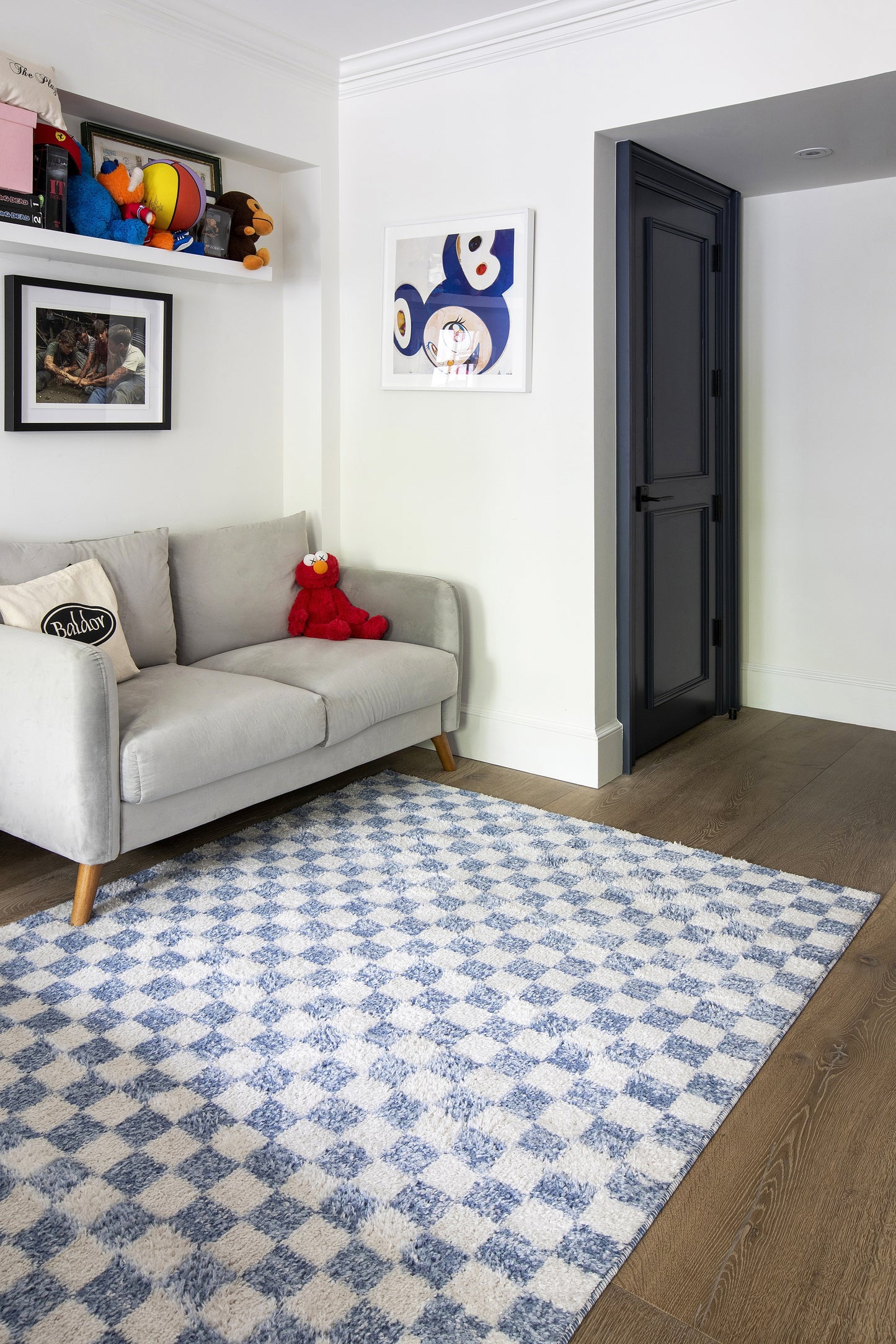 Modern living room: light gray fabric sofa, blue-and-white diamond rug, dark blue door, shelves with toys and art.